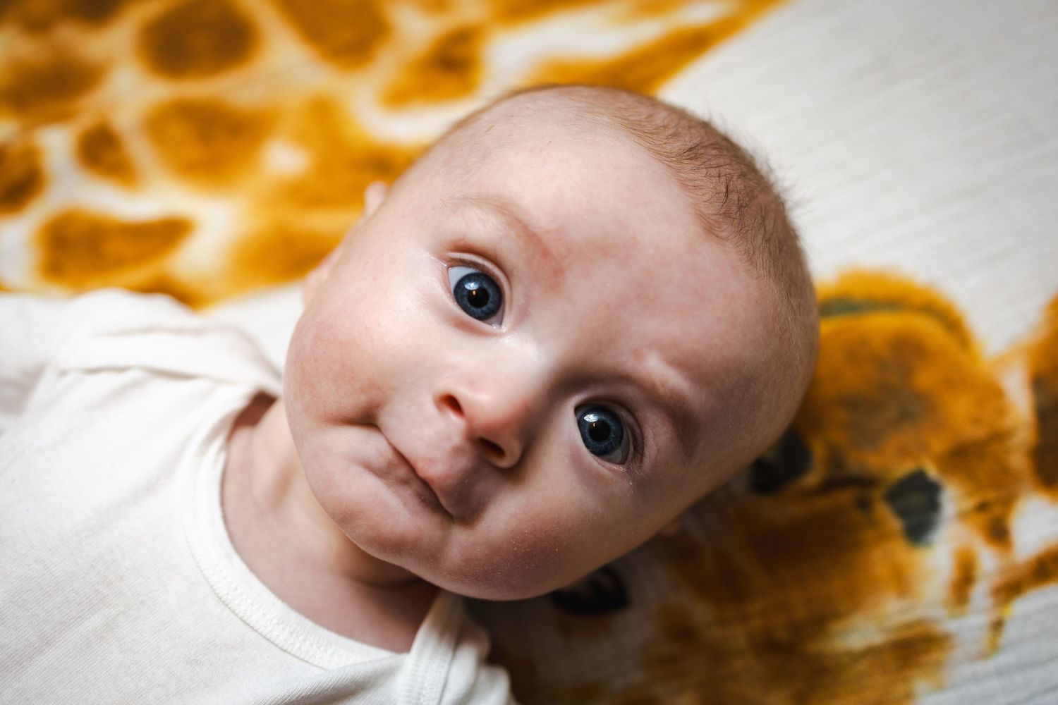 Baby wearing white clothing looks at camera with wide eyes against orange giraffe print background.