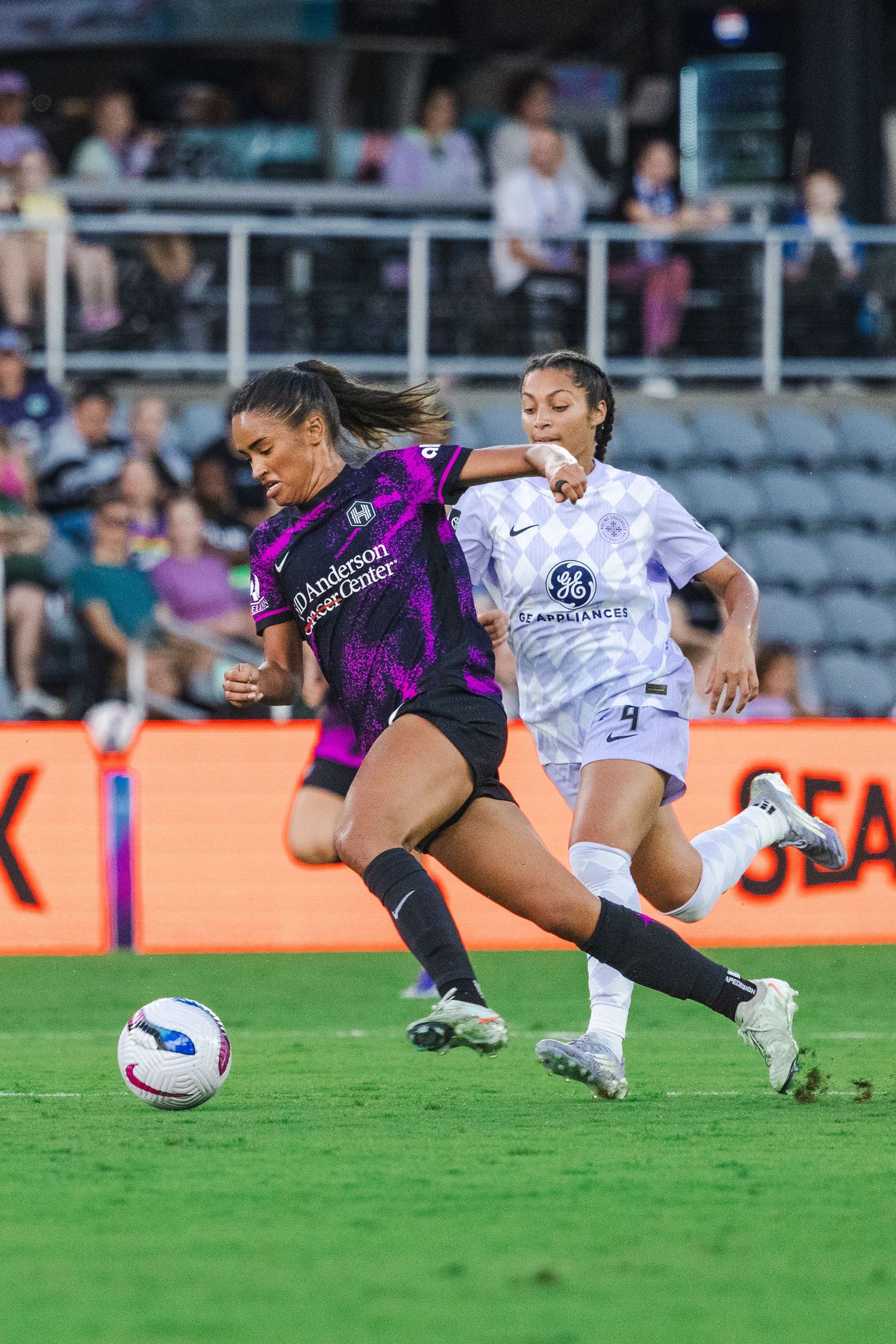 Two soccer players compete for the ball during a professional women's match between teams in purple and white uniforms.