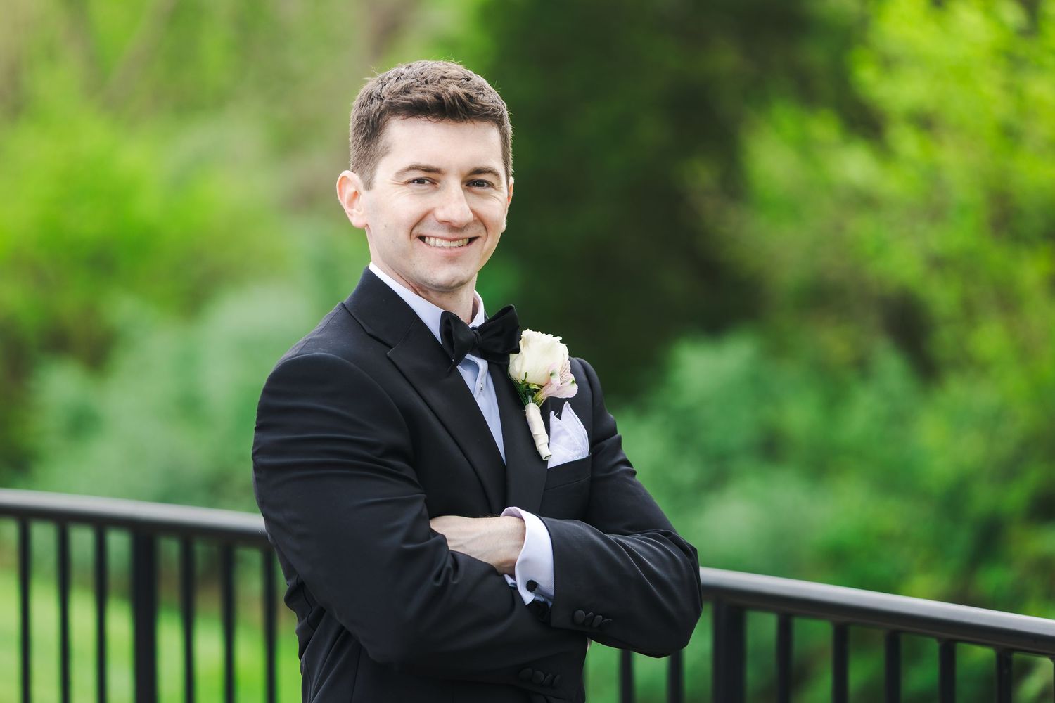 Smiling groom in black tuxedo with boutonniere standing outdoors with arms crossed.
