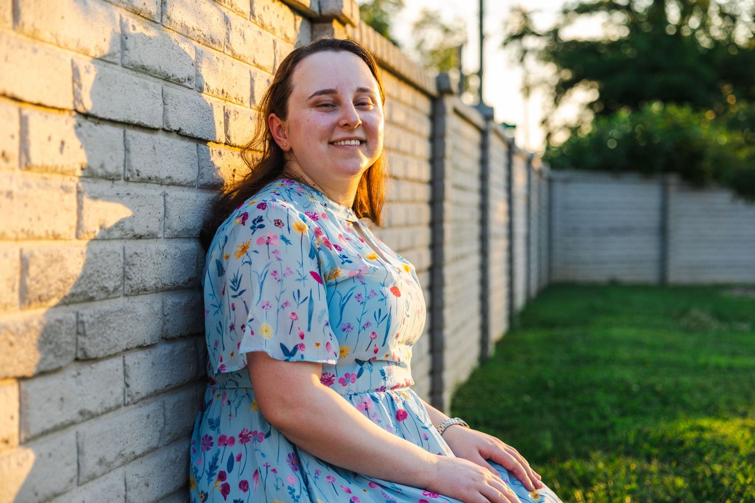 A person in a floral dress sits on a brick wall during golden hour lighting outdoors.