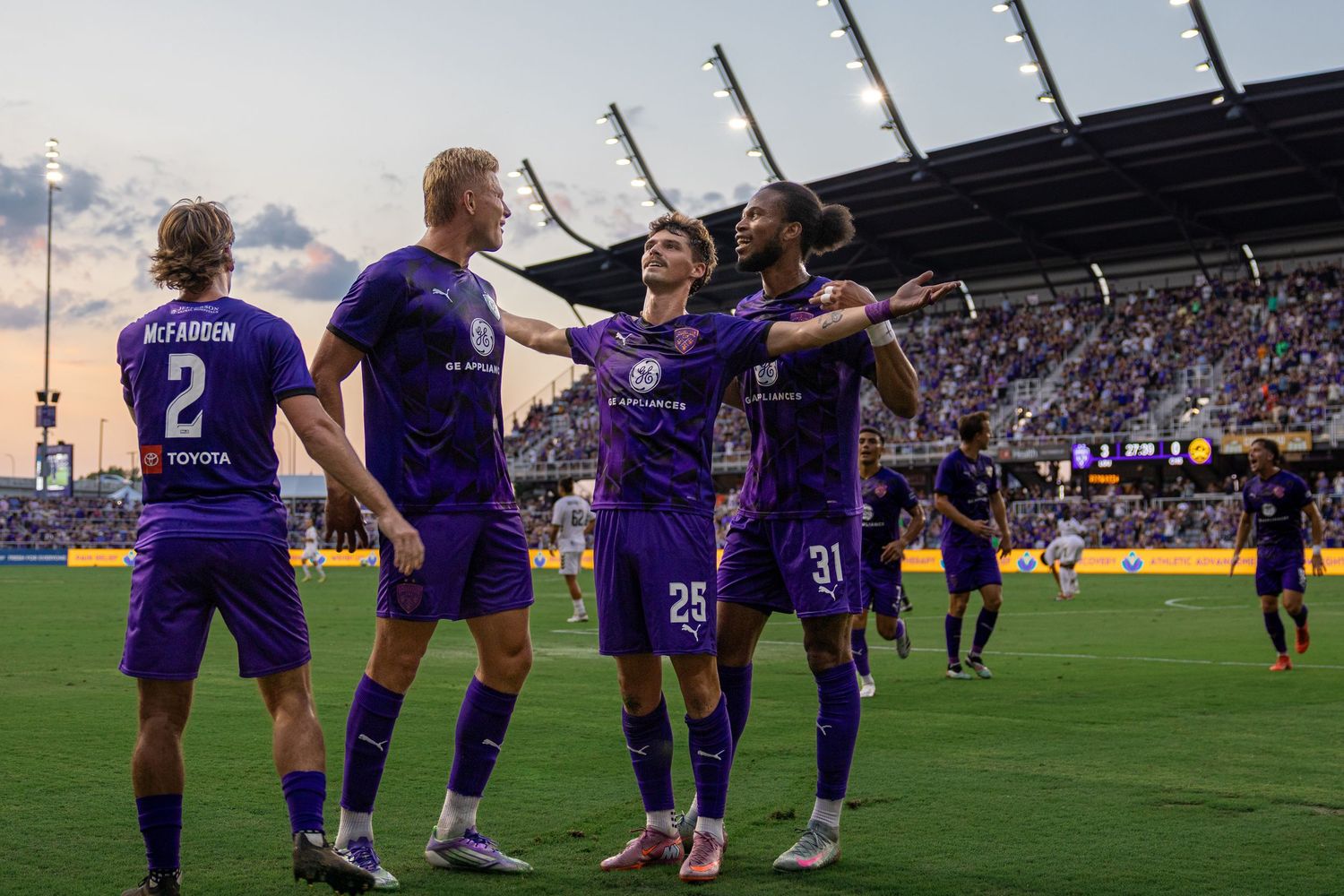Orlando City SC players in purple jerseys celebrate a goal together on the soccer field during an MLS match.
