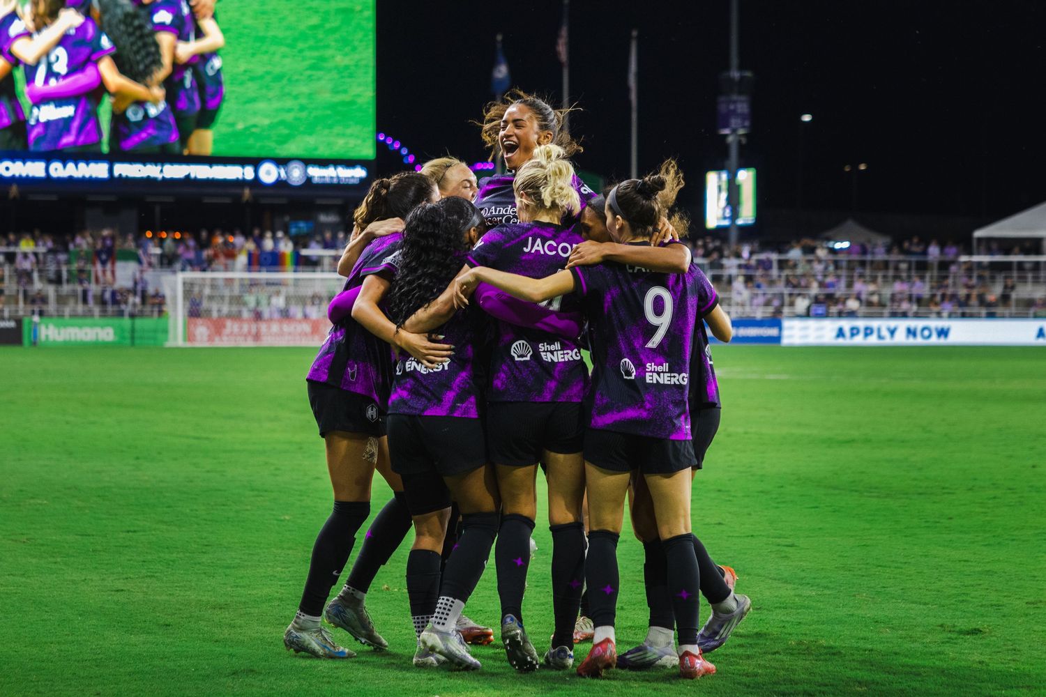 Women's soccer team celebrates on the field after scoring a goal in their purple uniforms.