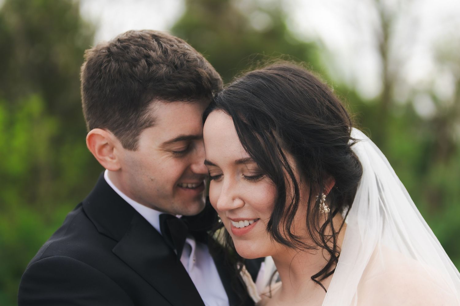 Bride and groom share a romantic forehead touch, smiling softly in an outdoor wedding portrait.