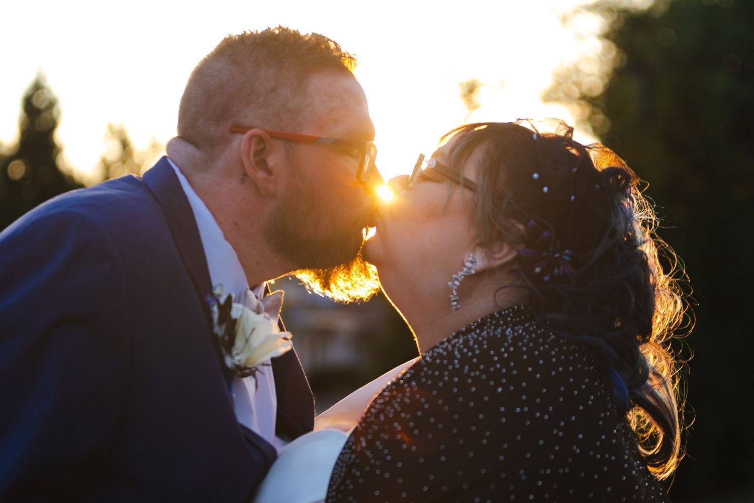 A couple shares a romantic kiss at sunset while standing in a garden setting.
