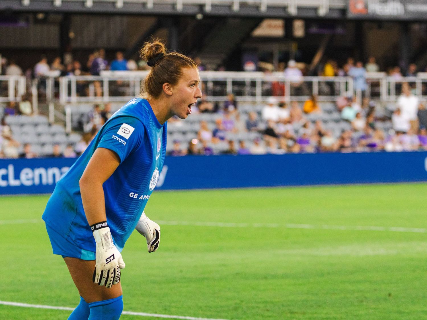 Soccer player in blue uniform stretches on field during pre-game warmup at stadium with spectators in background.