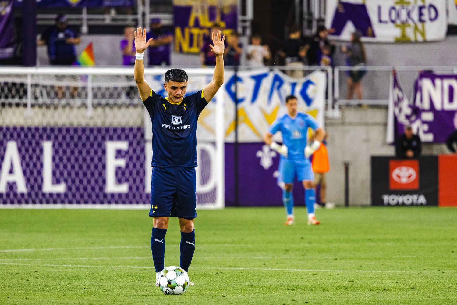 Soccer player in navy uniform raising arms while standing with ball on field.