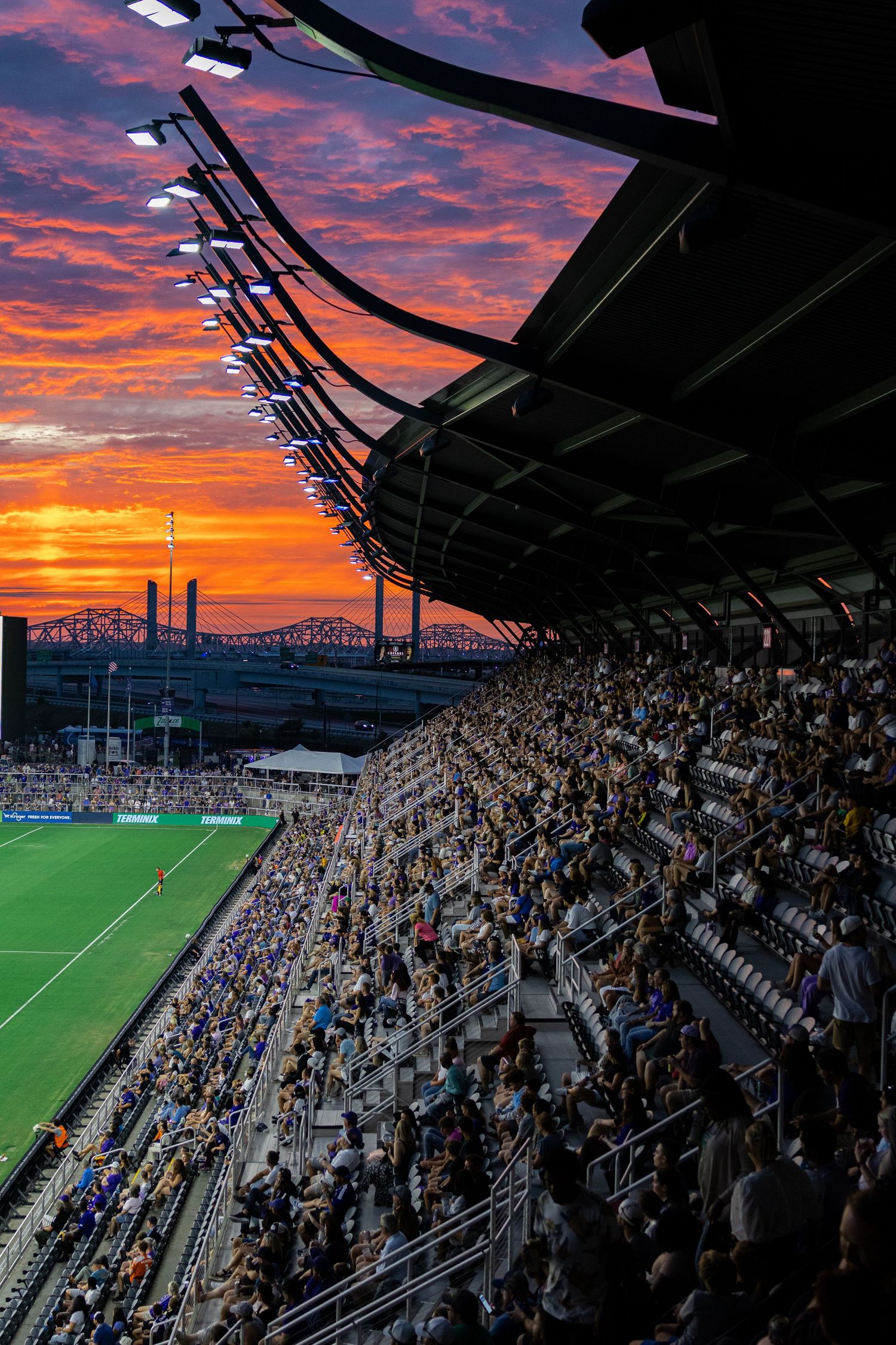 Dramatic sunset sky illuminates a crowded stadium with spectators watching a sporting event under modern roof architecture.