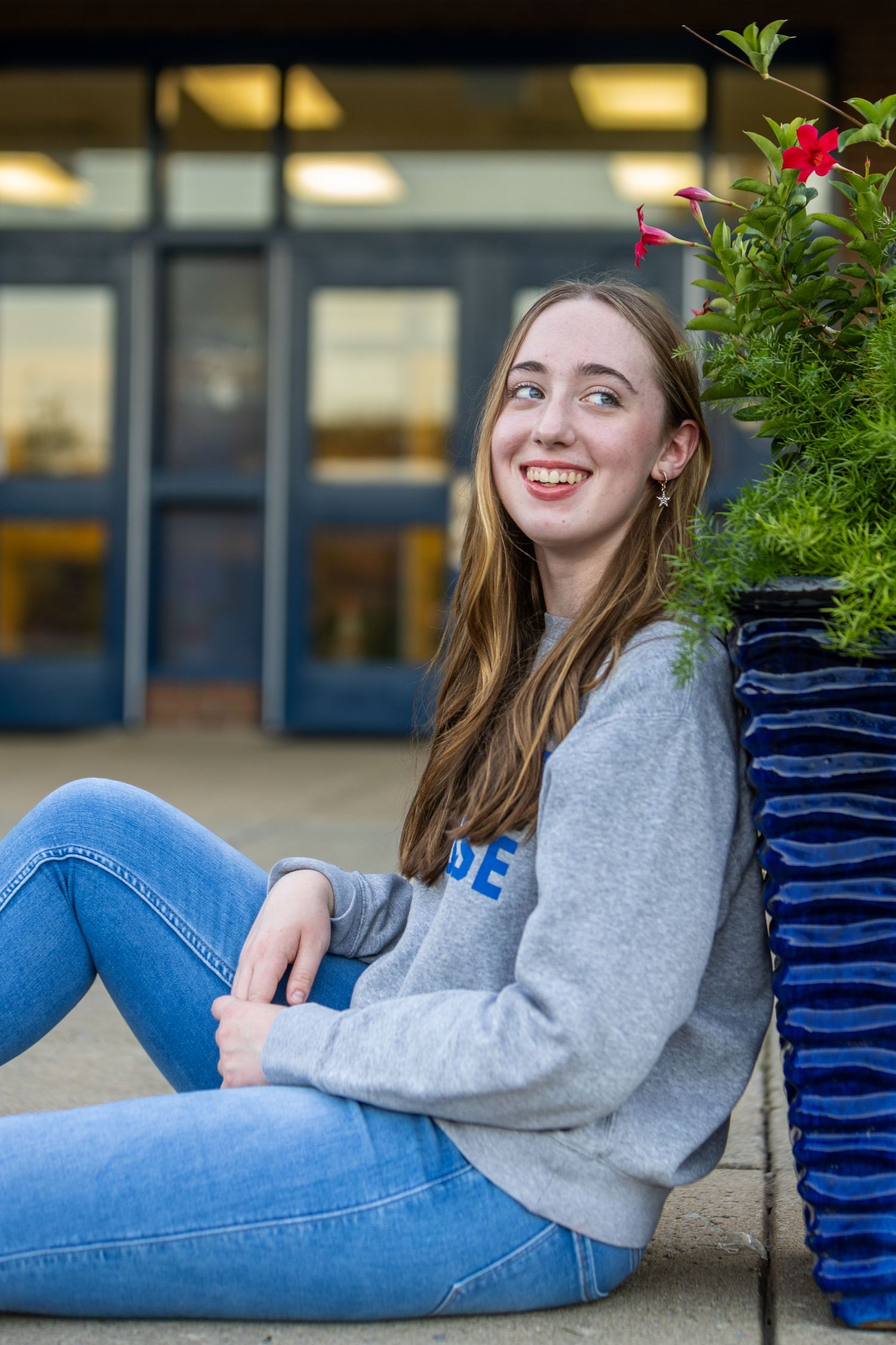 Student in grey sweatshirt and jeans sits casually outdoors near plants.