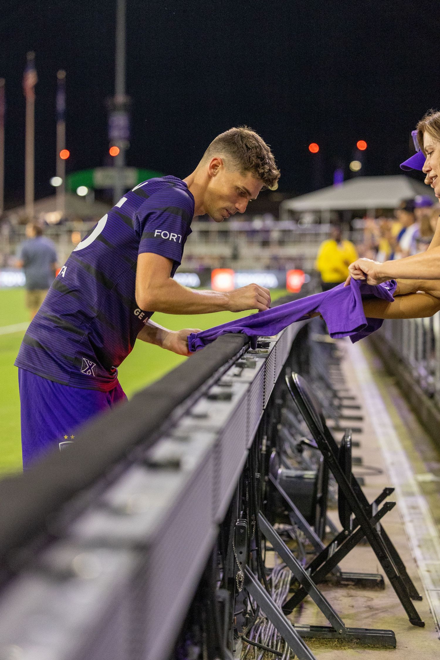 Soccer player signs autographs for fans at stadium railing after match.