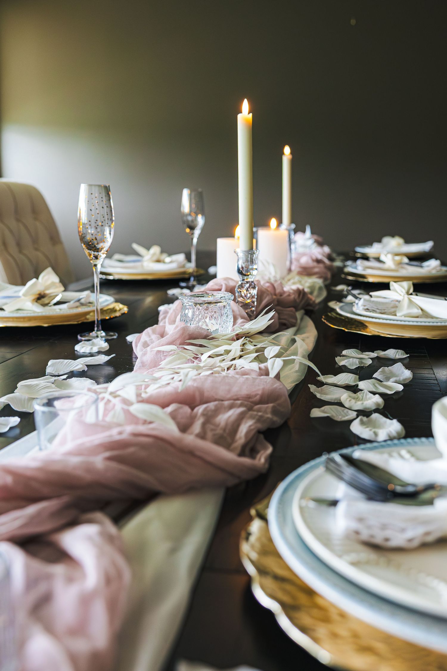 Elegant wedding table setting with pink gauze runner, white candles, gold charger plates, and scattered flower petals.