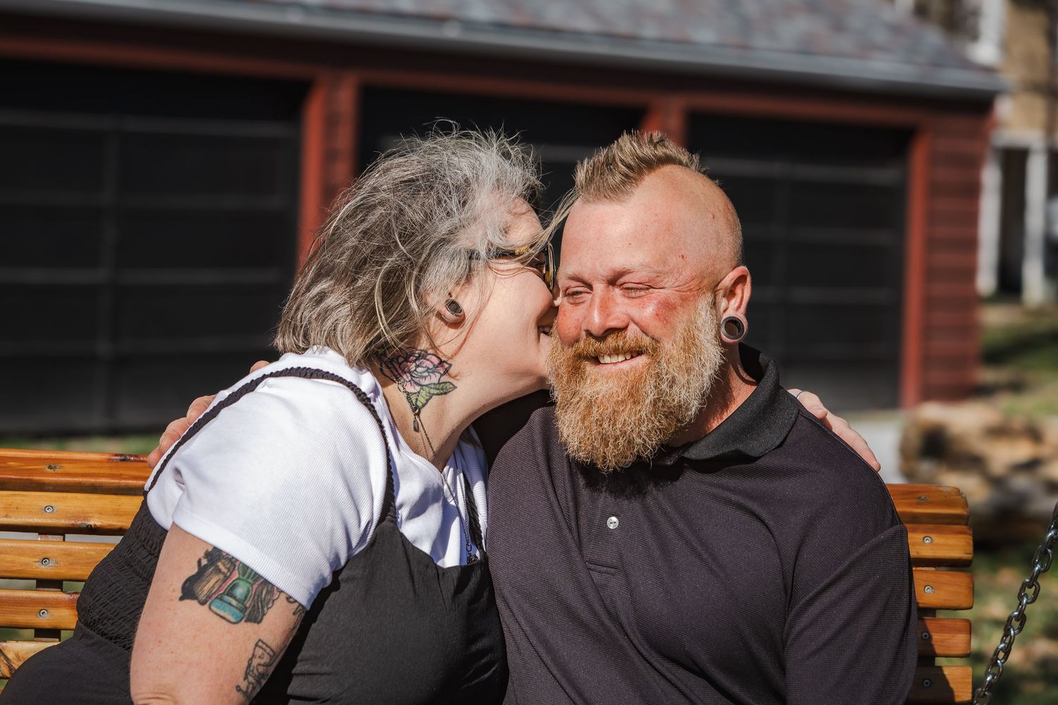Tattooed couple sharing an affectionate moment on a wooden bench.