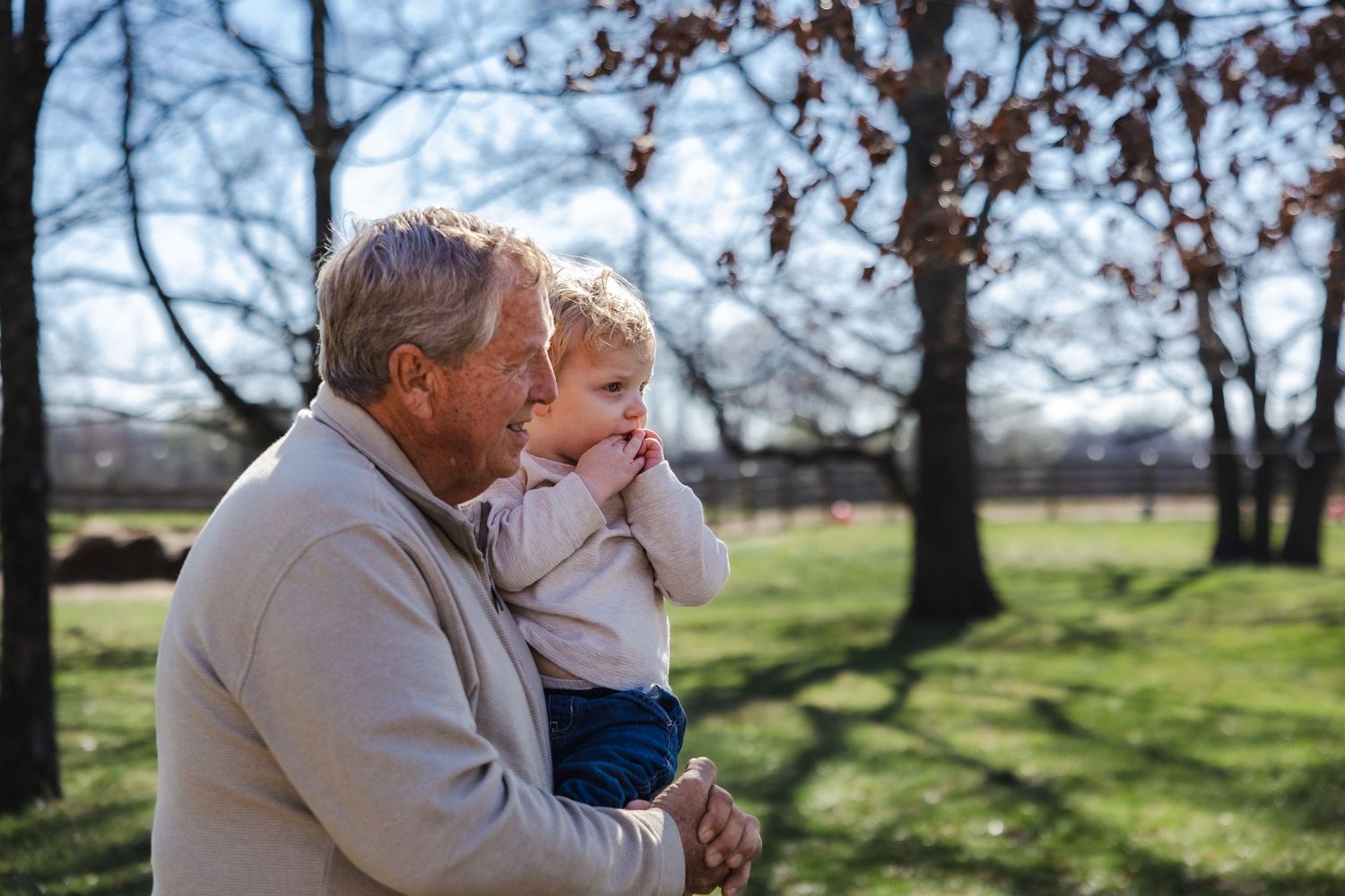 Two figures share a tender moment outdoors in a park setting with sunlit trees and green grass in the background.