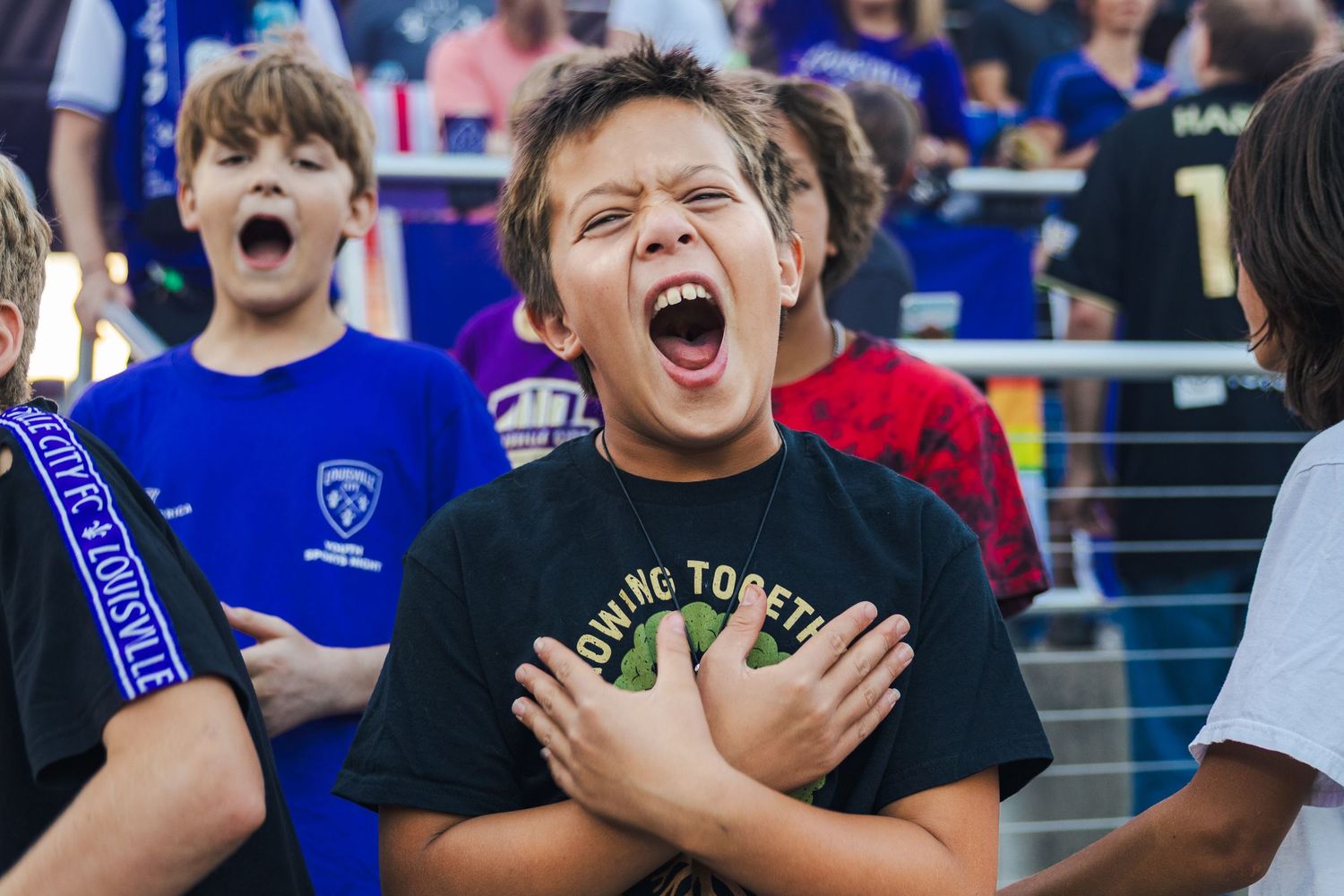 Excited fans cheer from the stands during a sports event at a stadium with blue seats and railings.