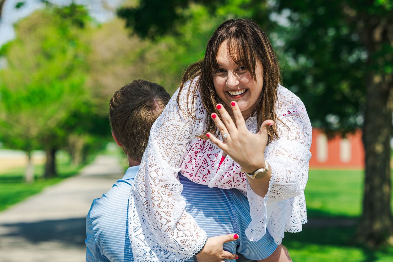 A joyful woman shows off her engagement ring while being lifted by her partner outdoors in a sunny park.