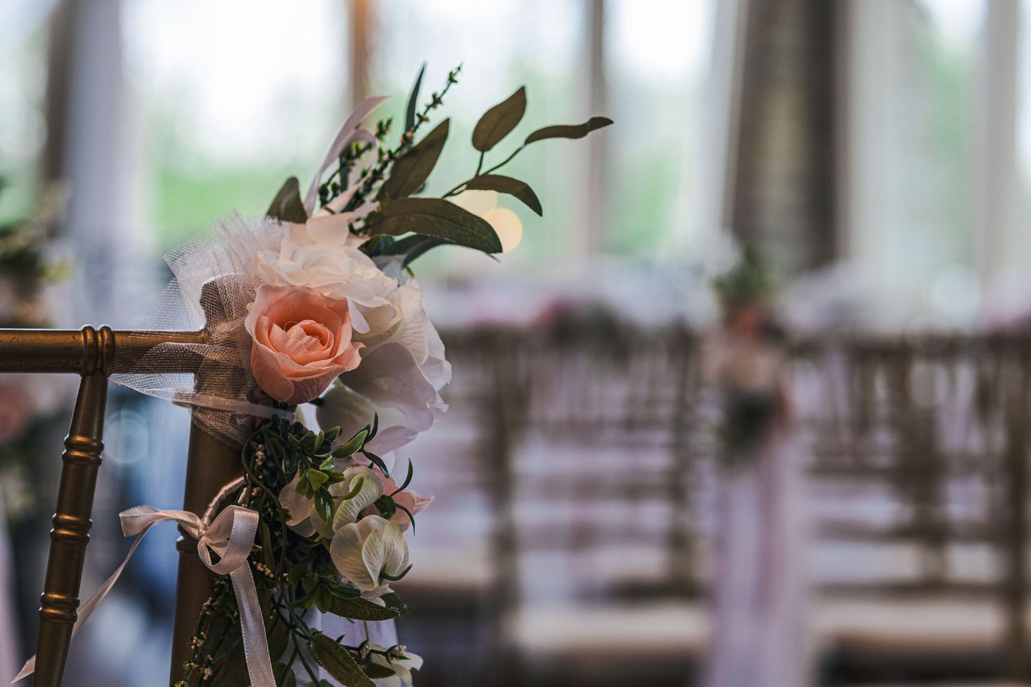 Elegant wedding aisle chair decorated with a blush pink rose, greenery, and white tulle ribbon at a reception venue.