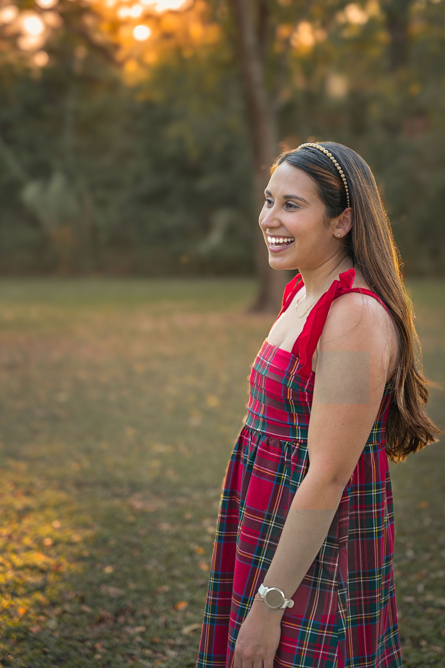 A person wearing a red and green plaid dress smiles while standing in an autumn outdoor setting at sunset.