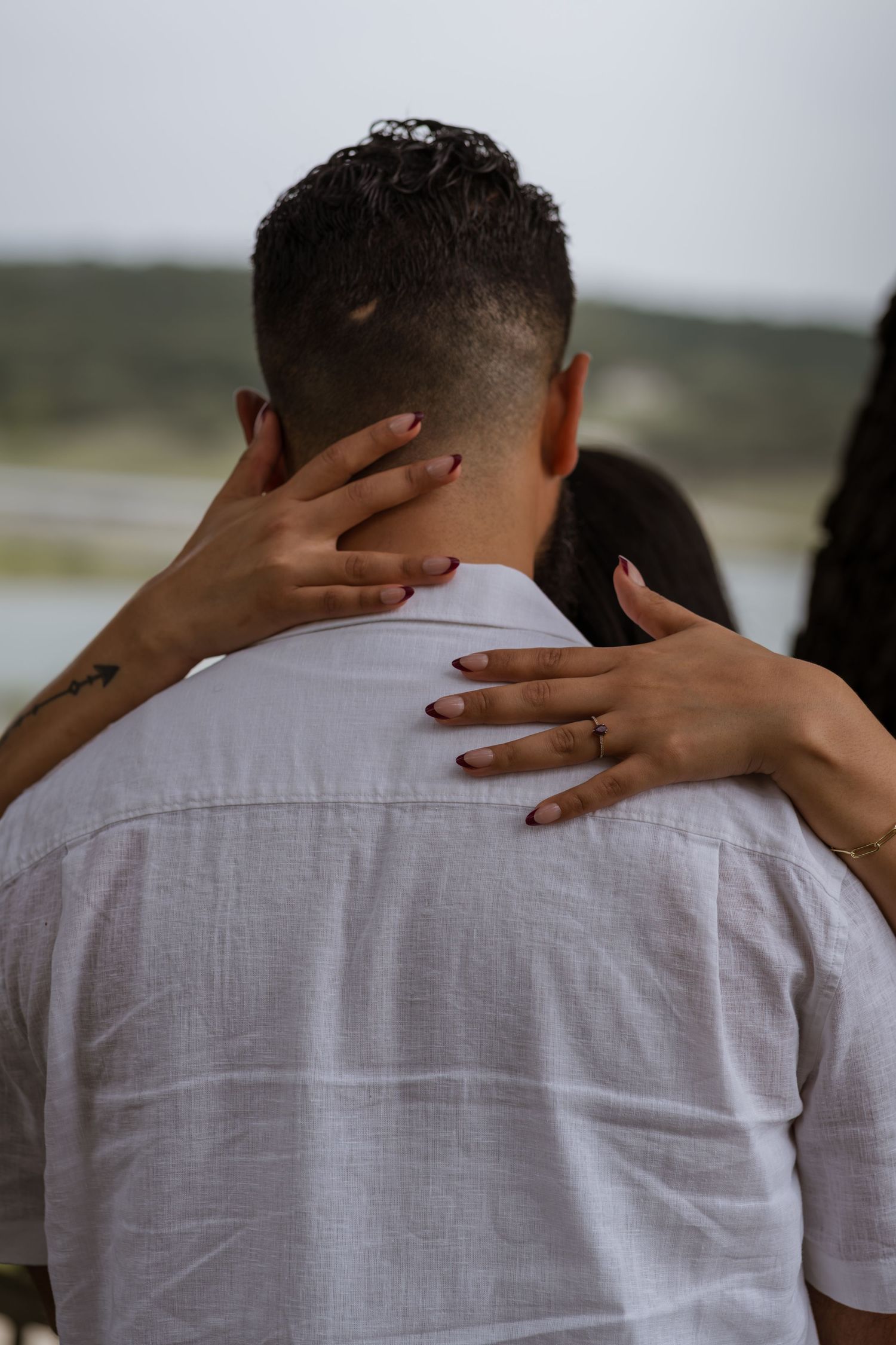 A woman embraces a man from behind, her hands resting on his shoulders, with a scenic outdoor backdrop.