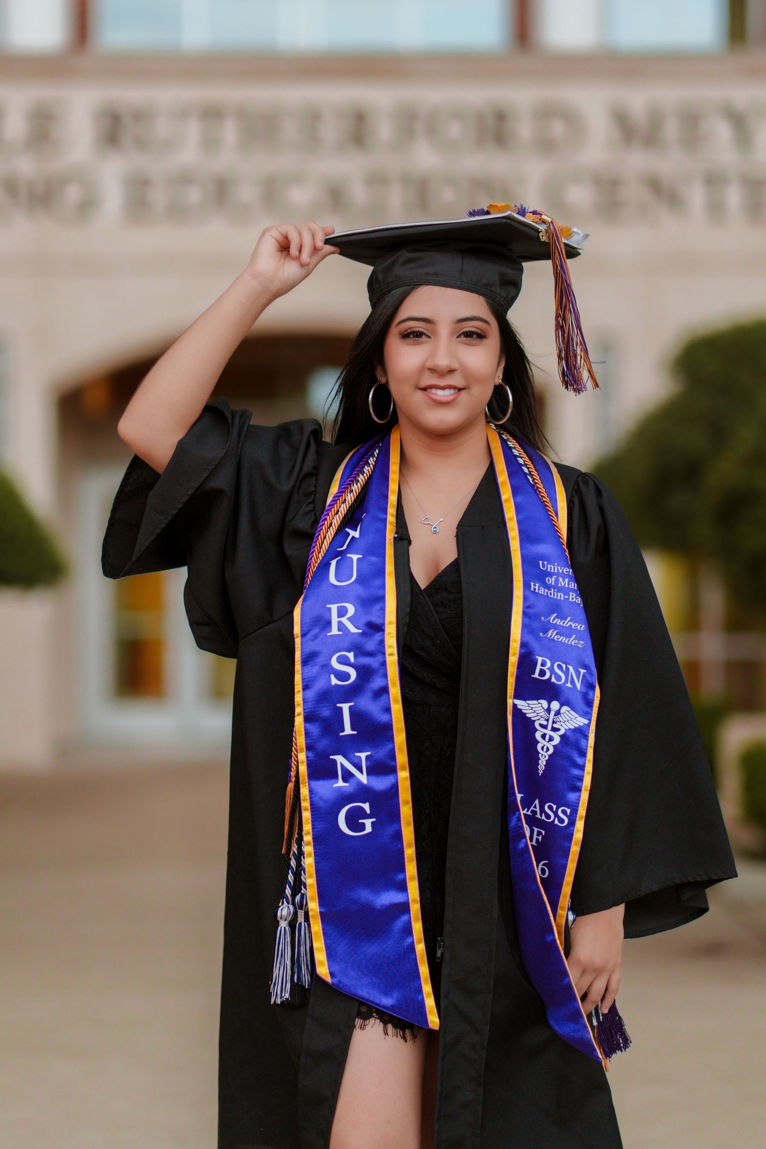 Nursing graduate in black cap and gown with BSN stole smiles outside education center.