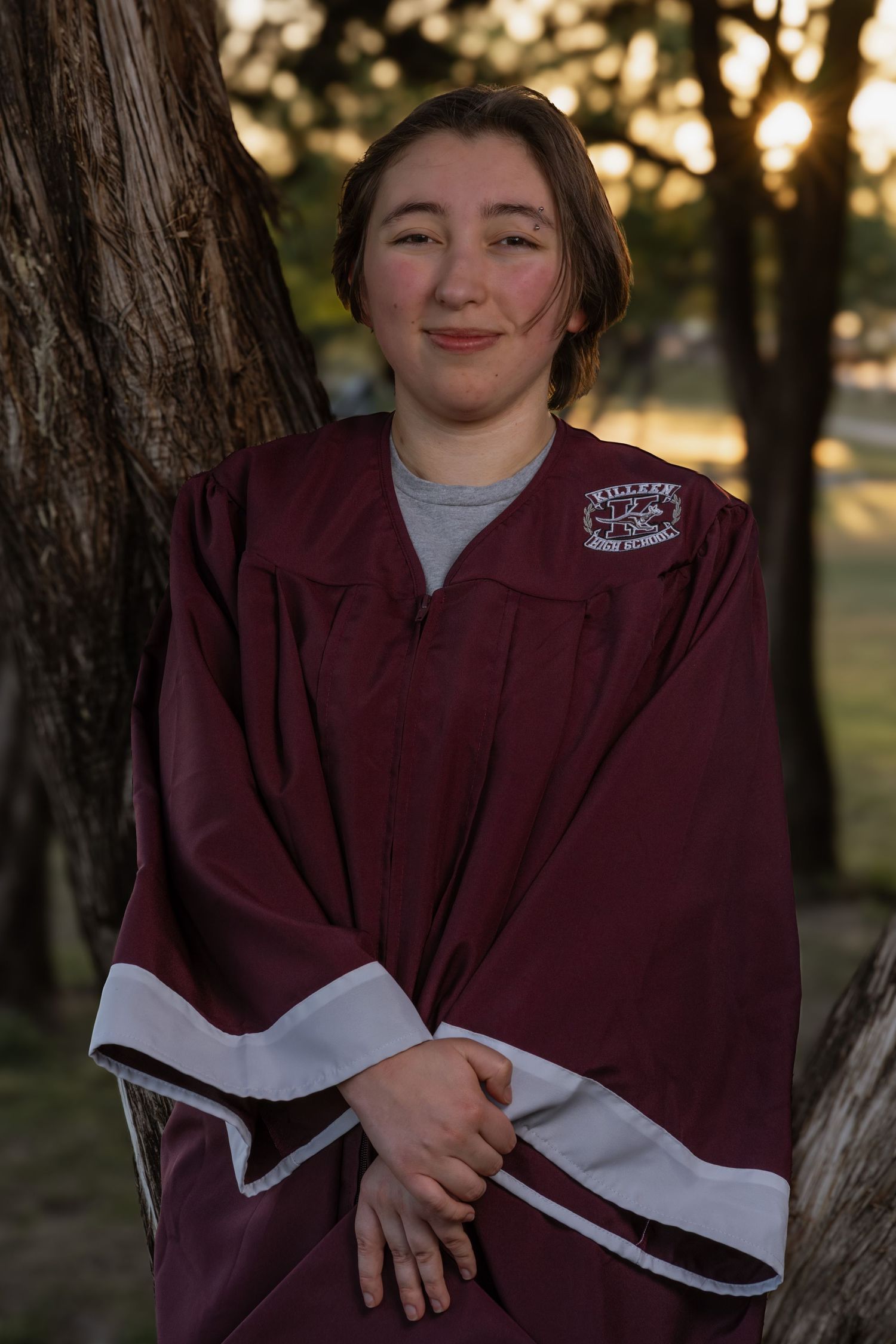 Young graduate smiling in maroon graduation gown, posing outdoors near a tree at sunset.
