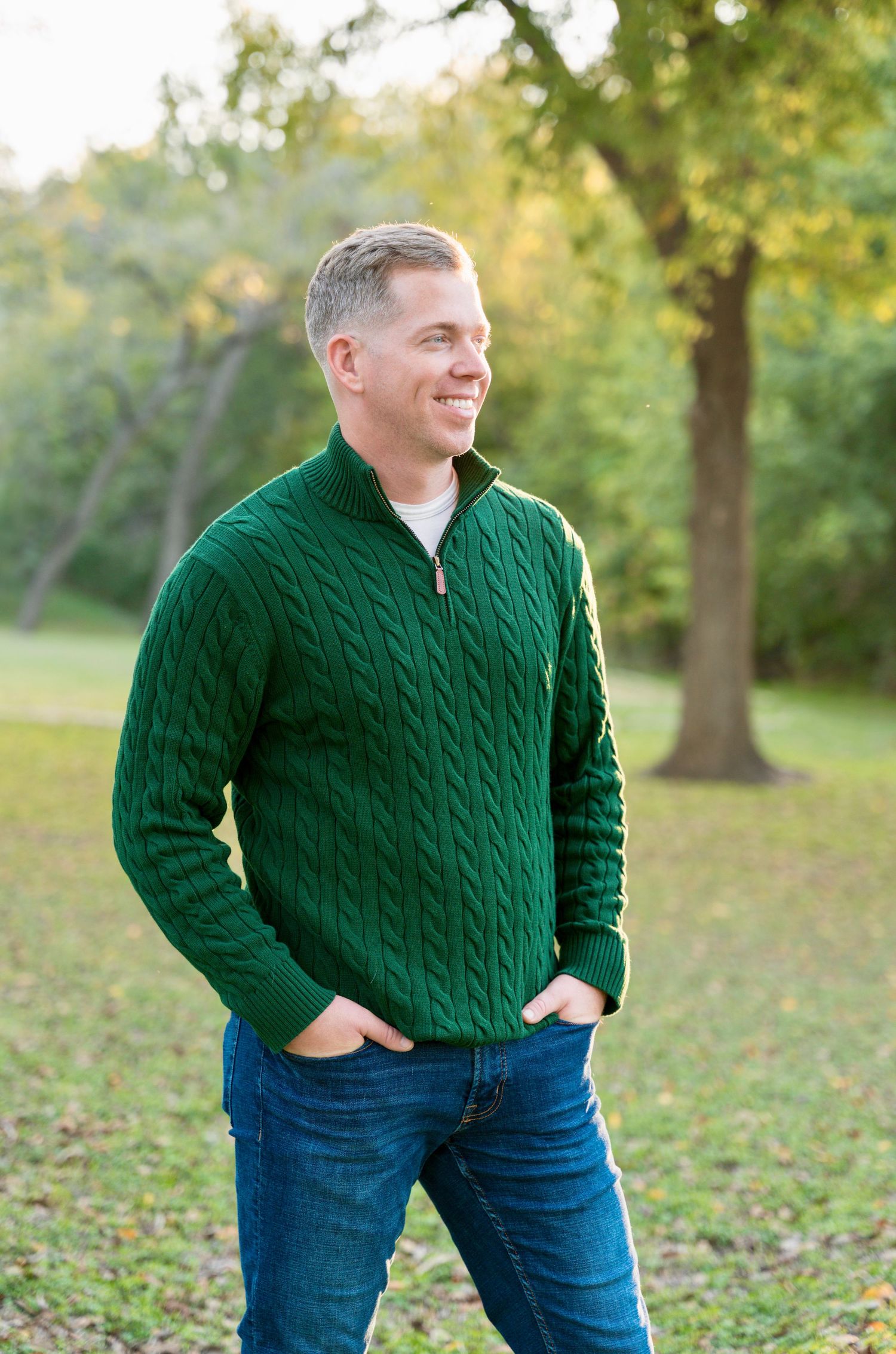 Man wearing a green sweater standing outdoors in a park during a natural light portrait session.