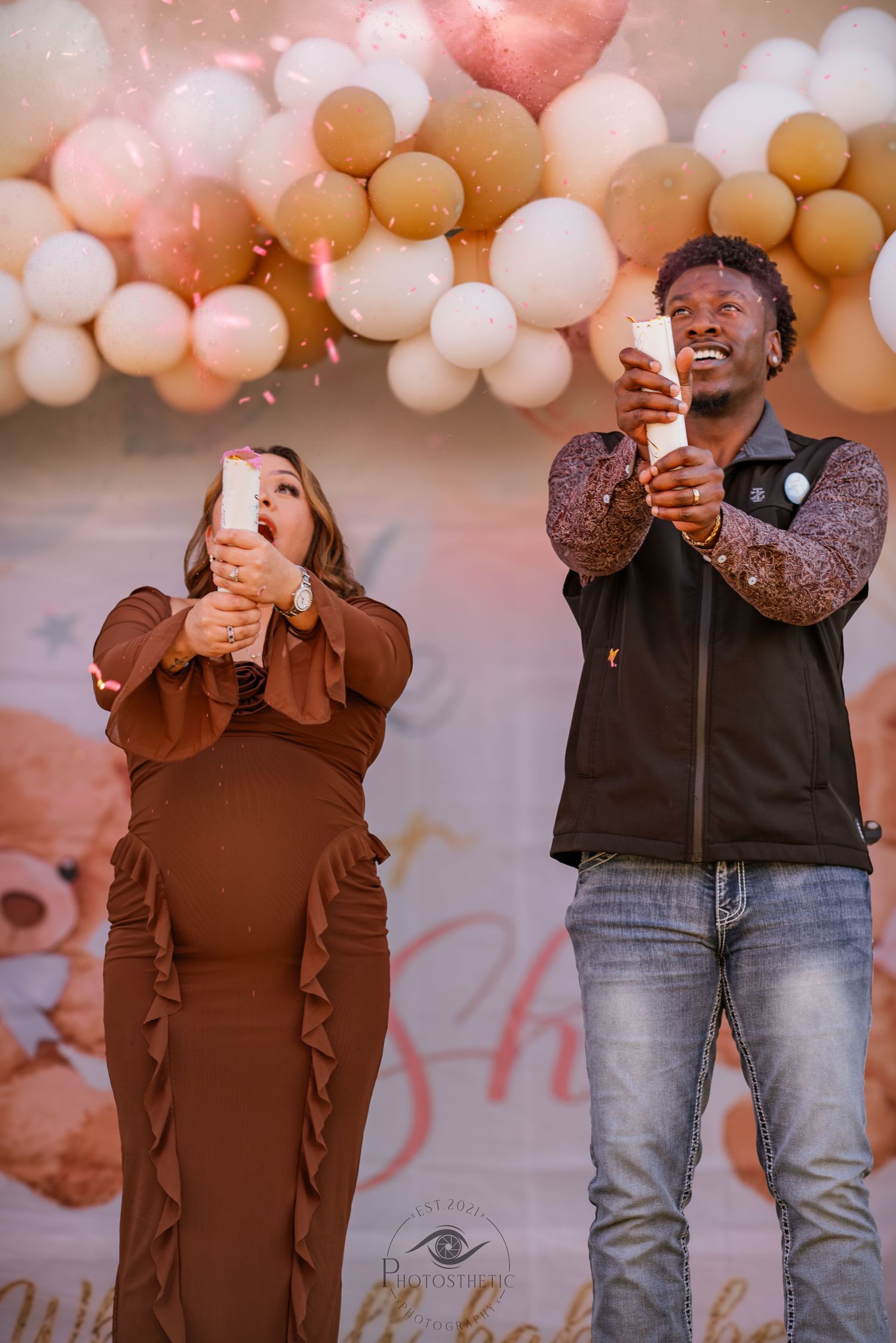 Two people laugh and celebrate in front of a peach and gold balloon arch backdrop at a festive event.