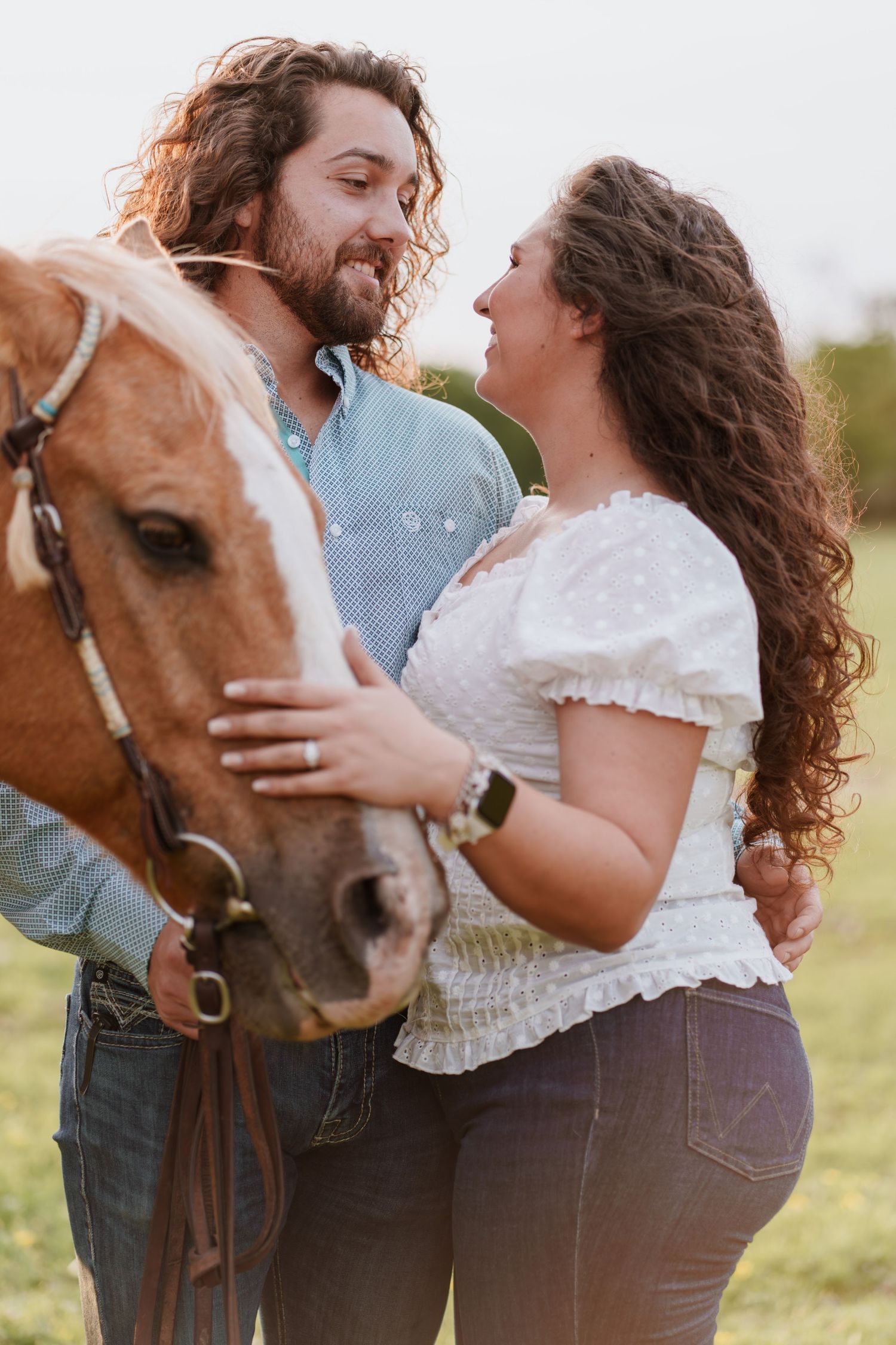 Couple smiling at each other while petting a chestnut horse in a sunny field.