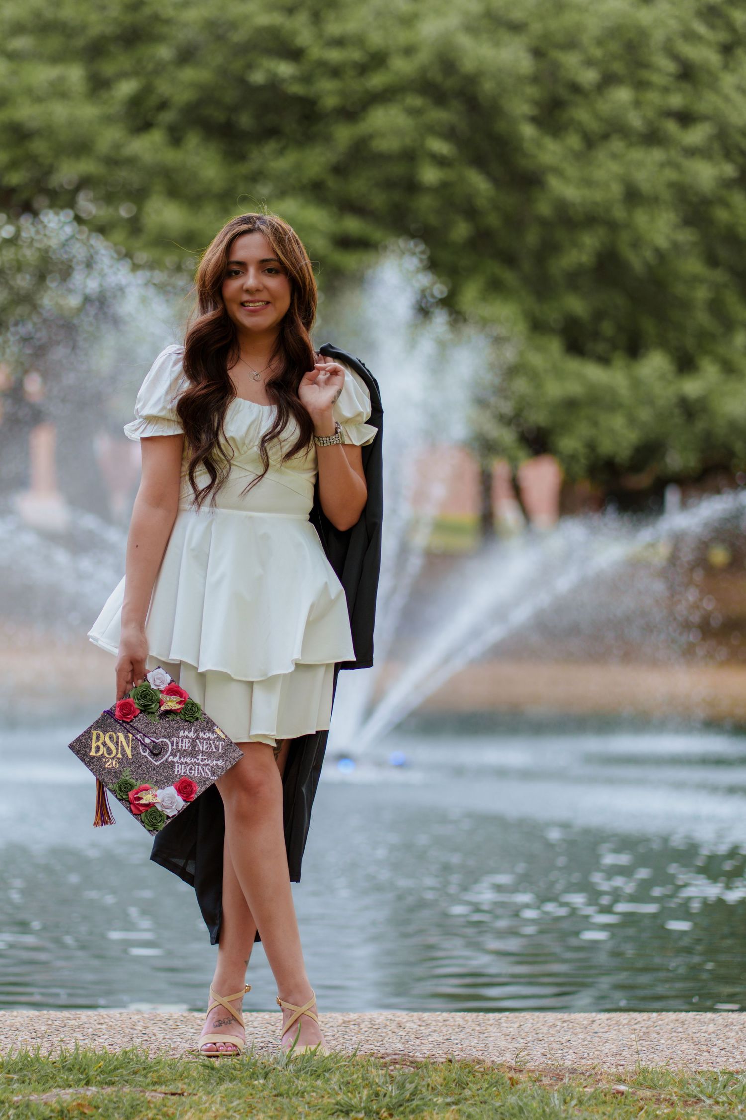 A smiling nursing graduate poses in a white dress holding her decorated BSN cap and gown near a fountain.