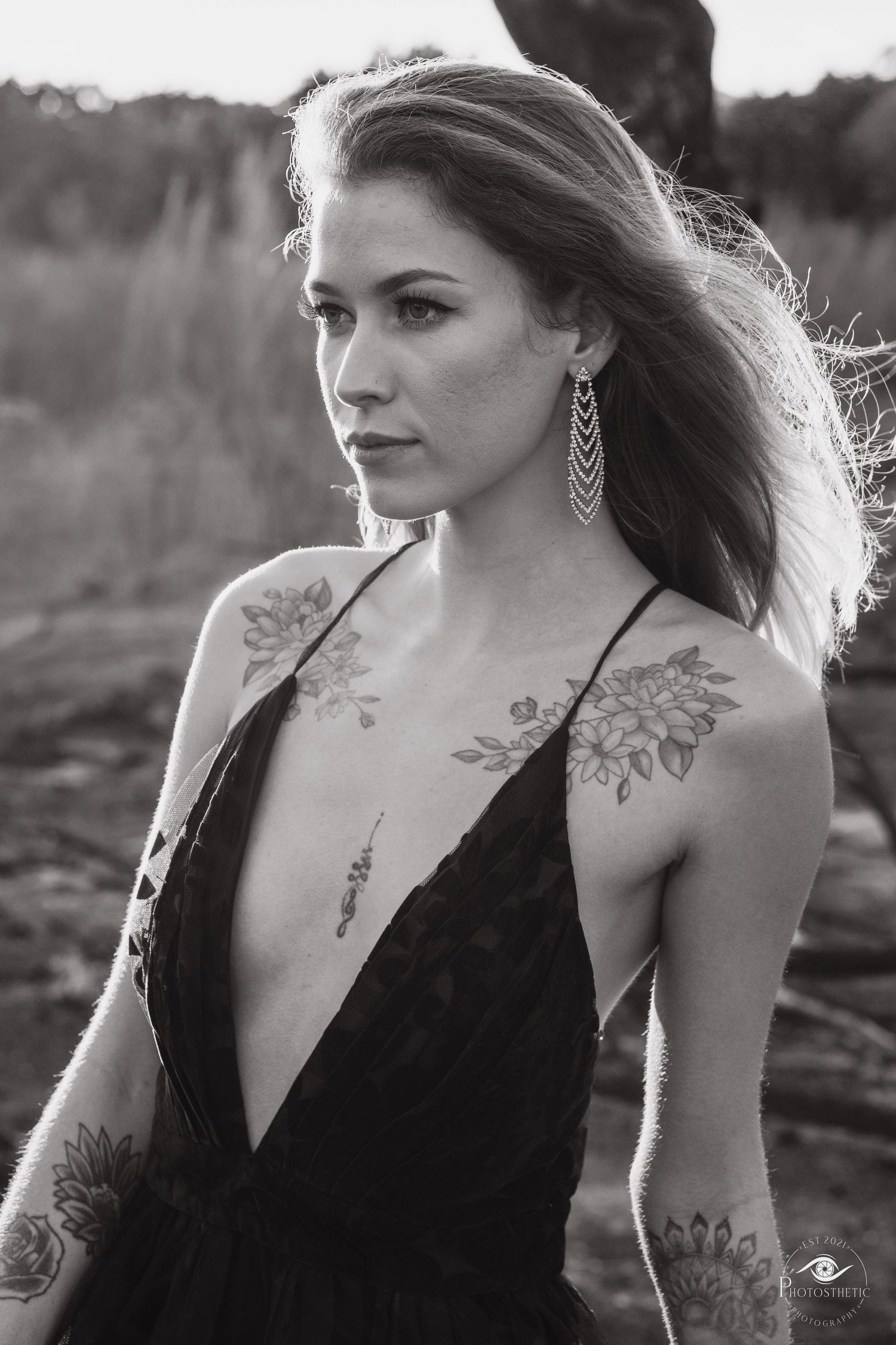 Black and white portrait of a woman with floral tattoos wearing a low-cut dress outdoors, looking thoughtfully into the distance.