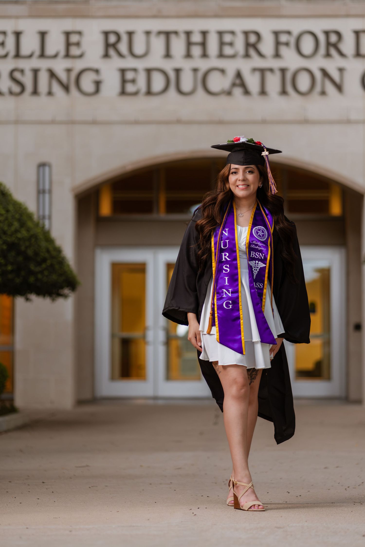 A nursing graduate poses in cap and gown with purple stole outside the Nursing Education Center.
