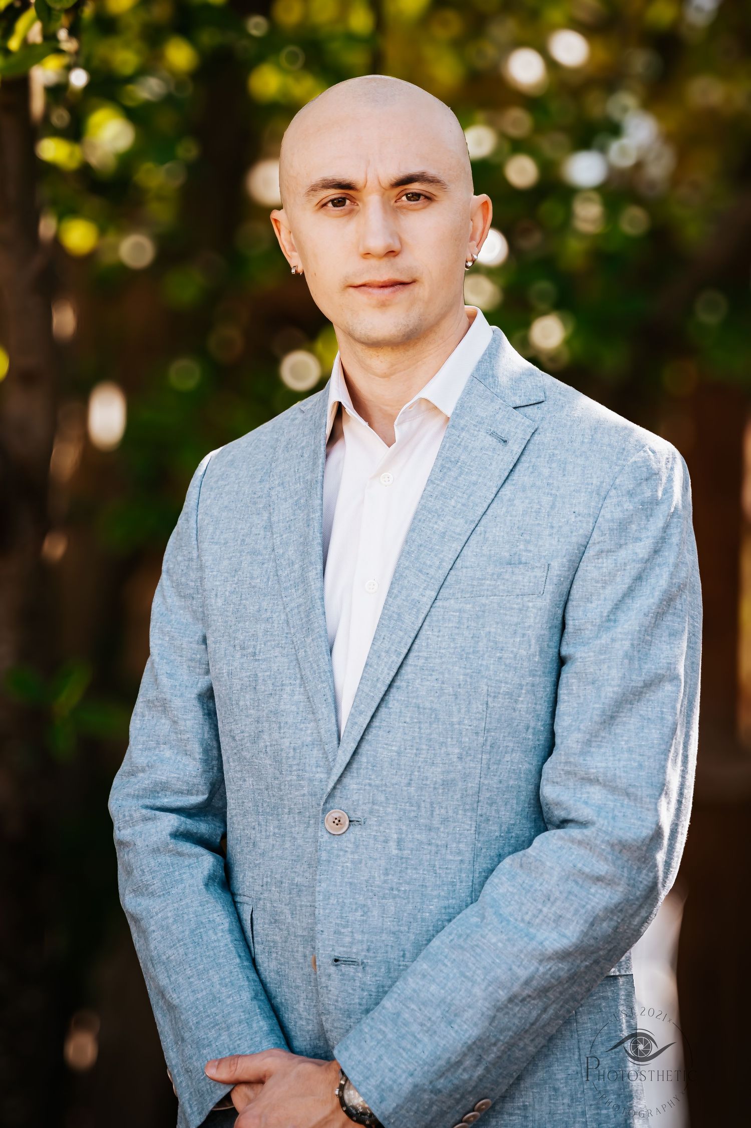 A person in a light blue suit jacket and white shirt poses outdoors against a blurred natural background.