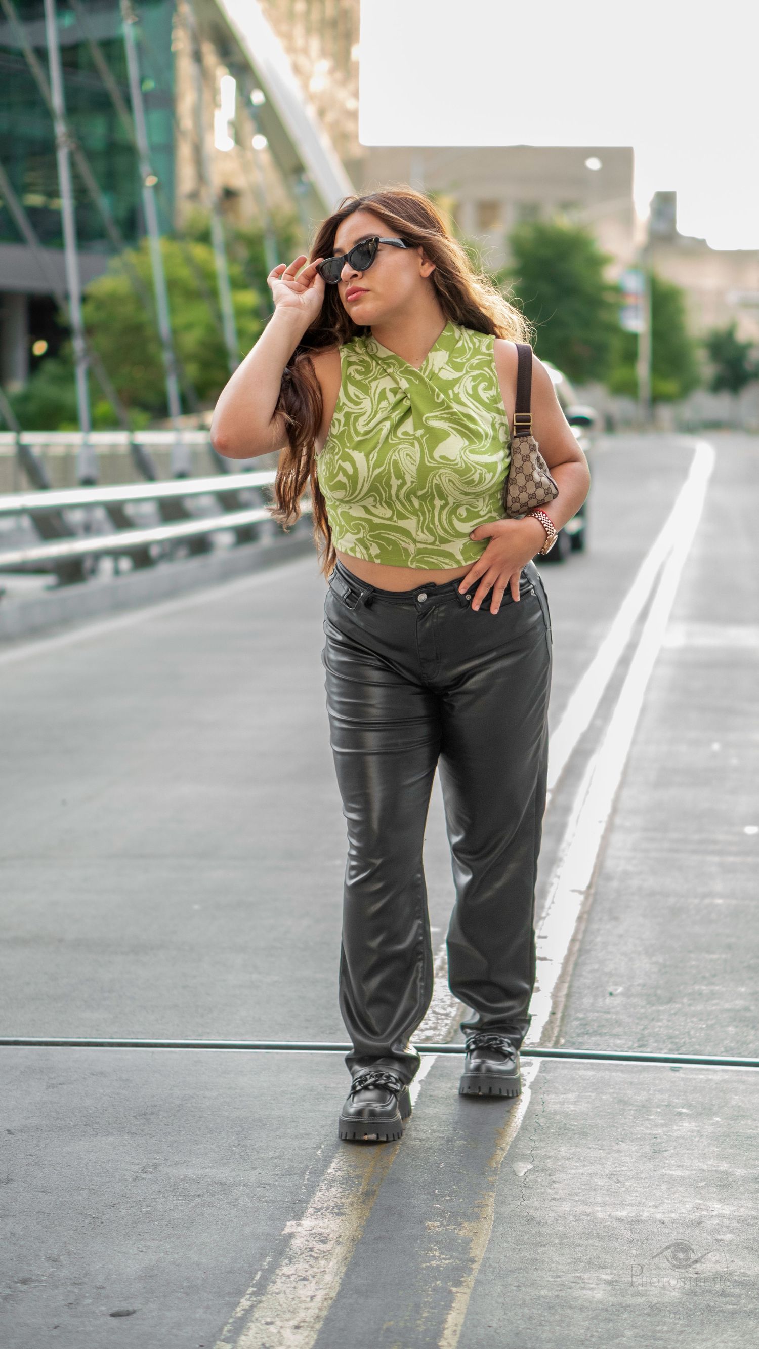 Woman wearing a green patterned top and black pants walking confidently on a city bridge.