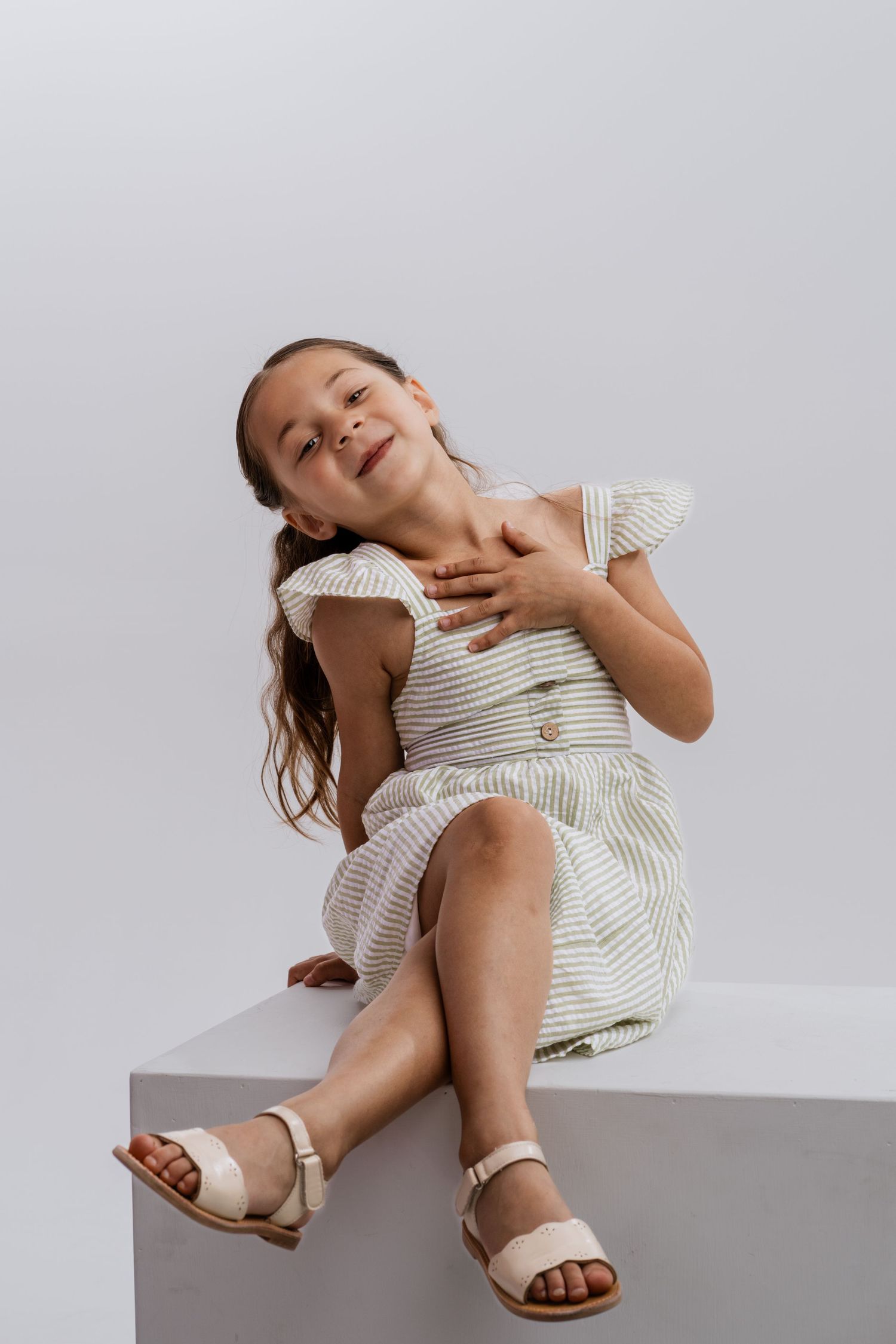 Young girl in striped dress and sandals sitting on white cube, smiling joyfully with hands on chest.