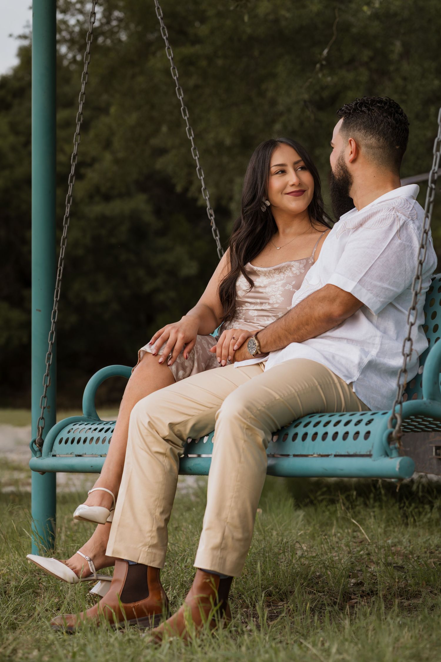 A couple sits together on a teal chain swing in a park, sharing a tender moment surrounded by lush green trees.