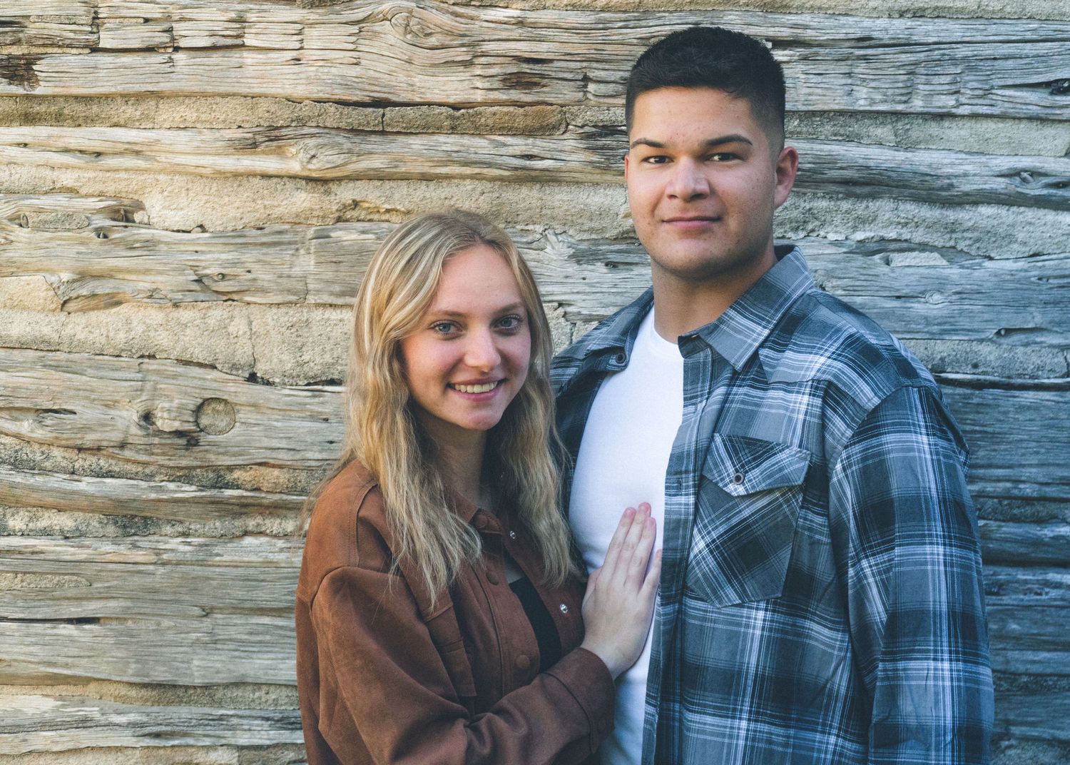 Couple standing together outdoors in front of a rustic wooden wall, smiling during a casual portrait session.