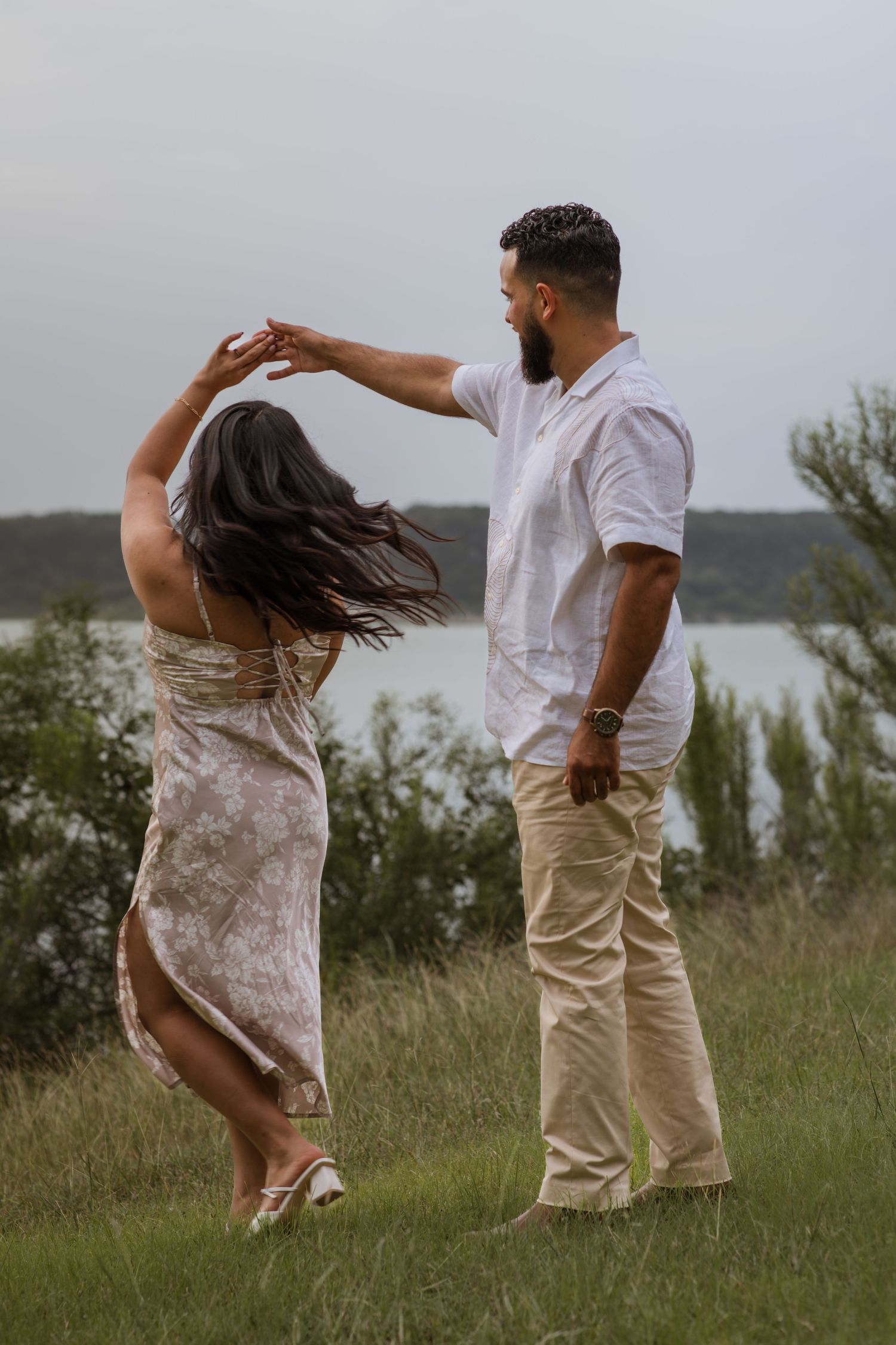 Couple dancing outdoors on green grass near a lake, man spinning woman in floral dress on cloudy day.