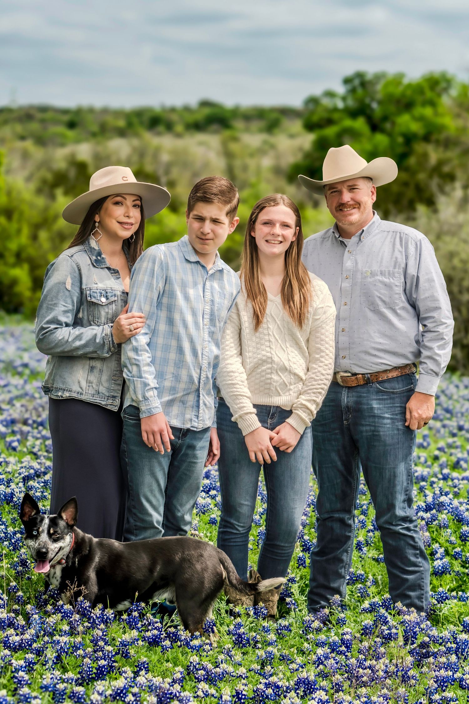 Family of four standing together in a bluebonnet field with their dog during a spring outdoor portrait session.