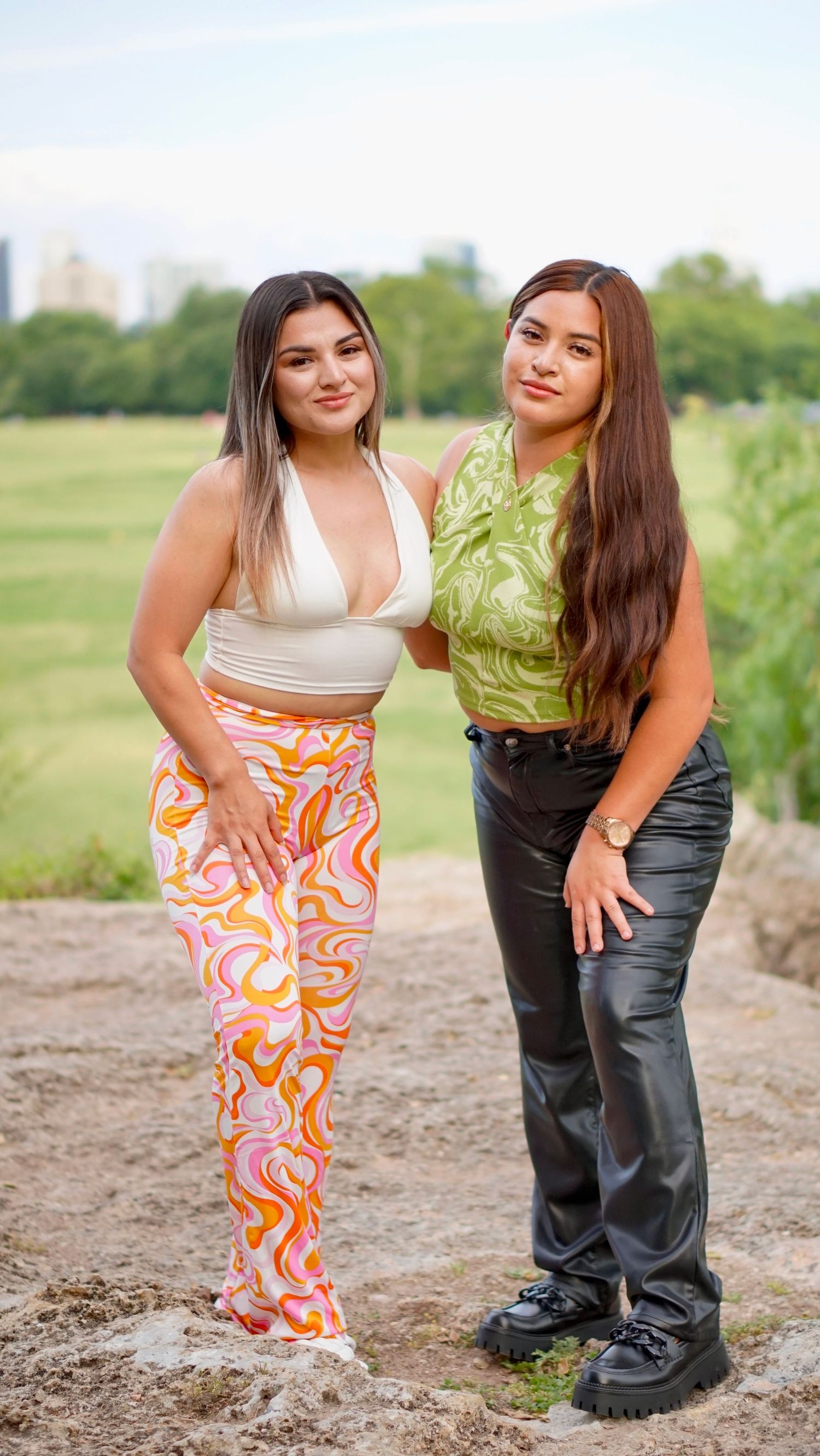 Two friends pose outdoors in summer outfits with a green park and Austin, Texas city skyline visible in the background.