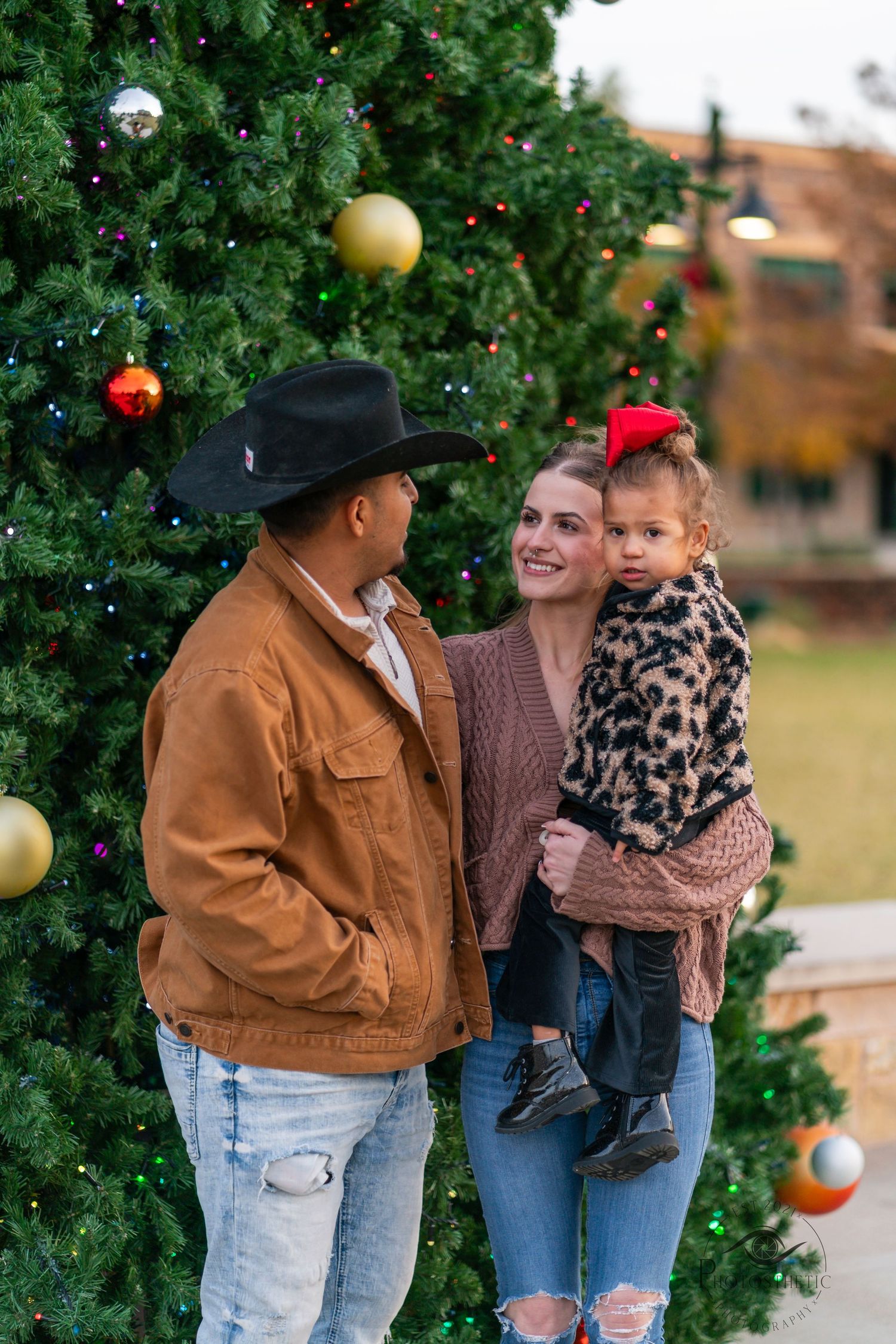 Family wearing cozy winter outfits poses together by decorated Christmas tree outdoors in casual holiday photoshoot.