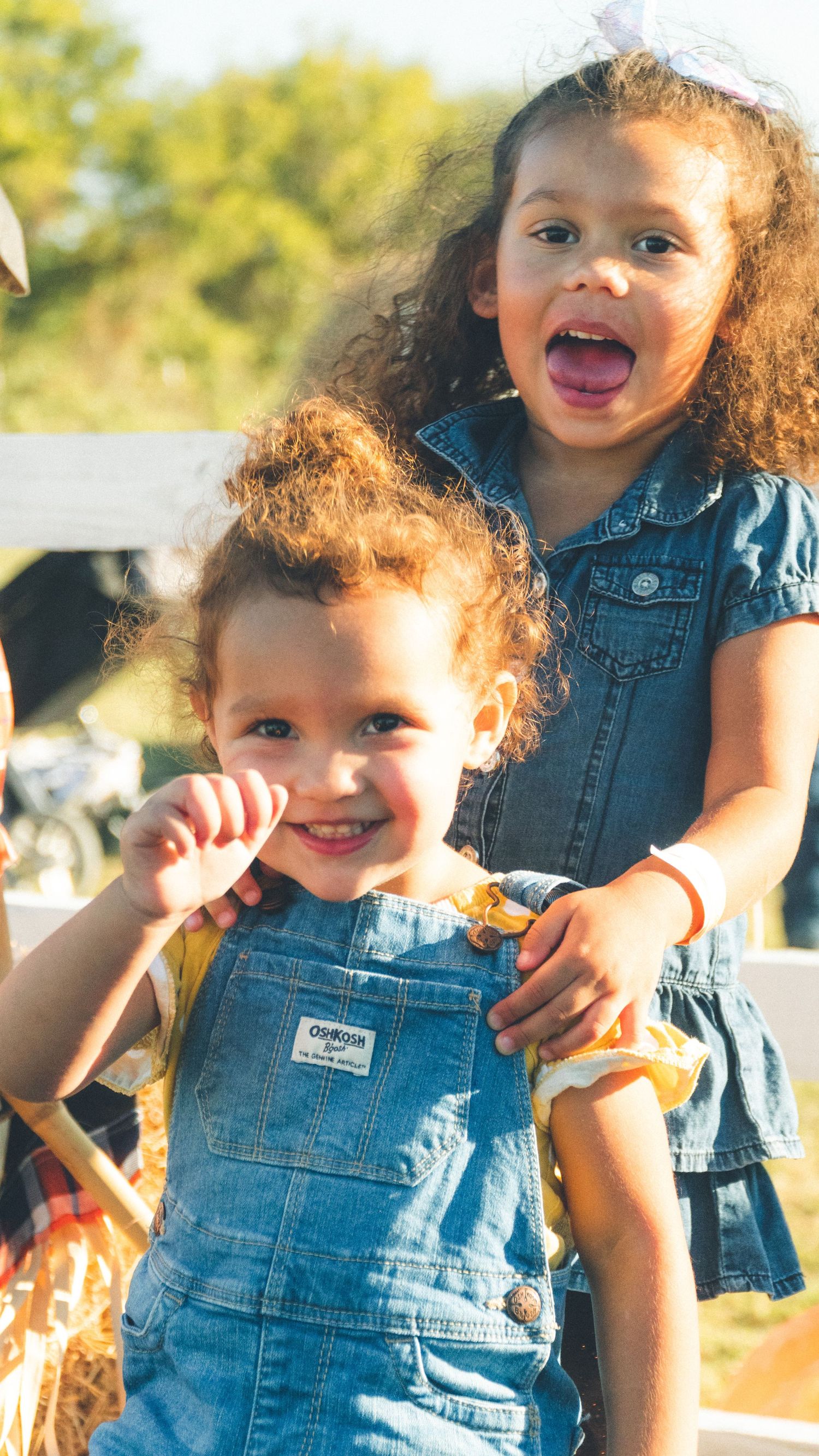 Two young children smiling outdoors, wearing denim outfits in natural light.