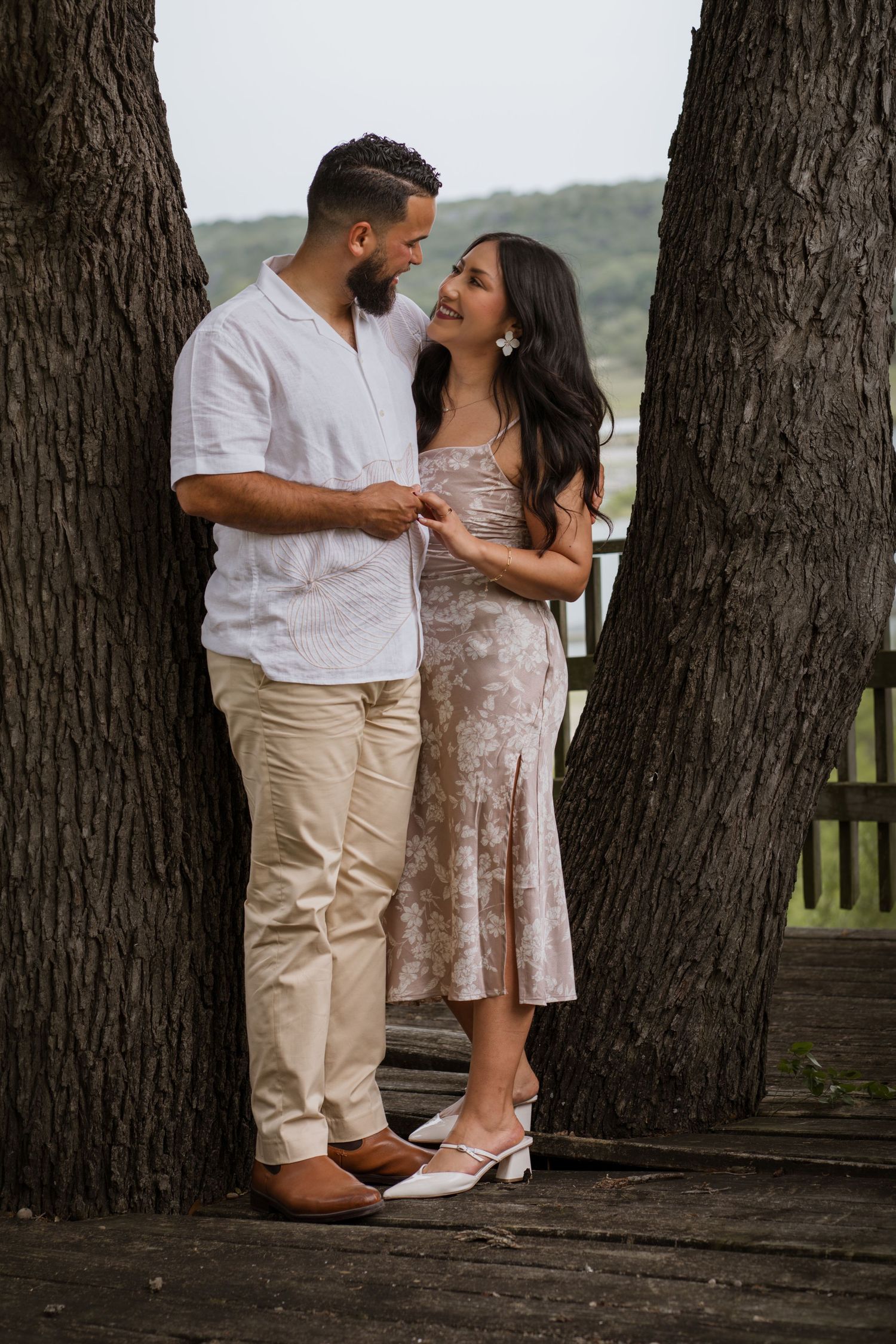 Couple sharing a romantic moment between two trees on a wooden deck, woman in floral dress, man in white shirt.