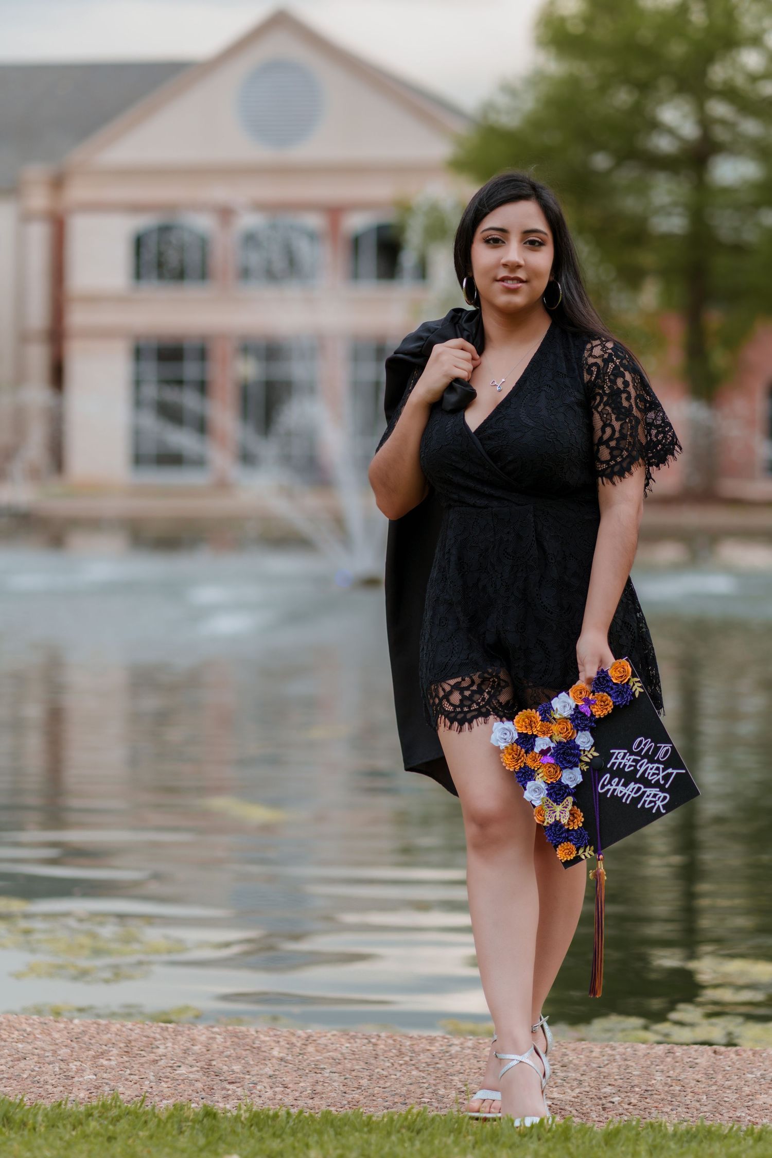 A young woman in a black lace dress holds a decorated graduation cap by a fountain, celebrating her academic achievement outdoors.