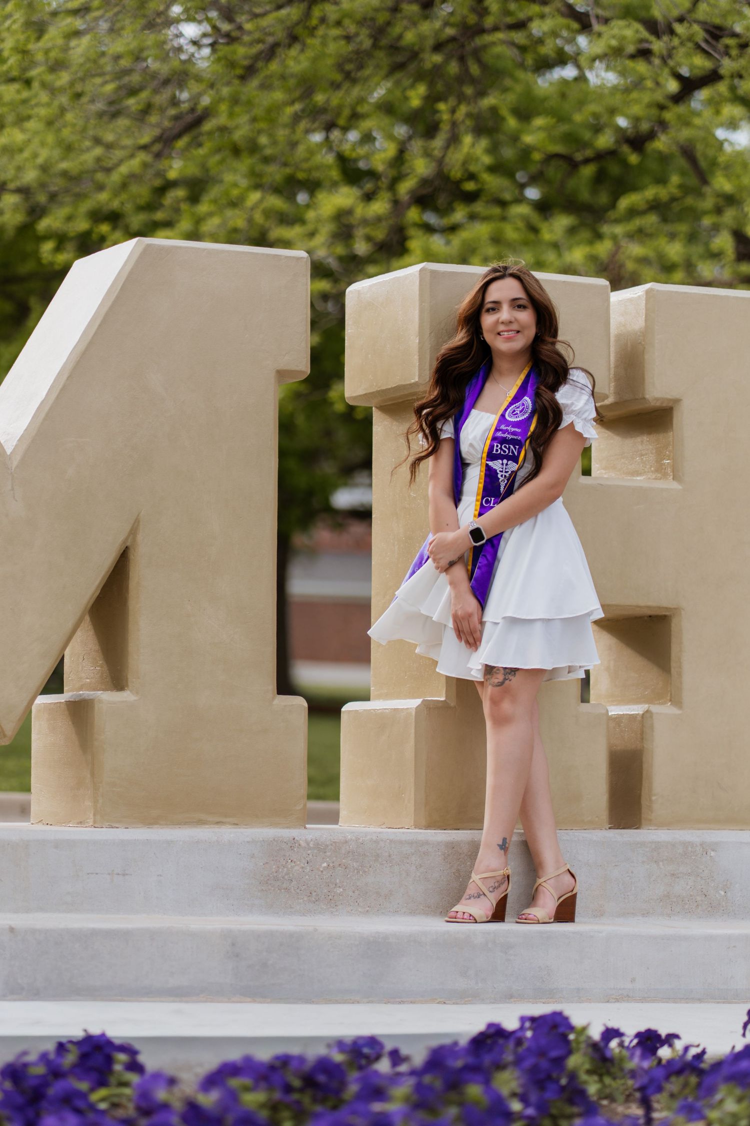 A smiling female graduate in a white dress and purple sash poses proudly in front of large stone letters on campus.