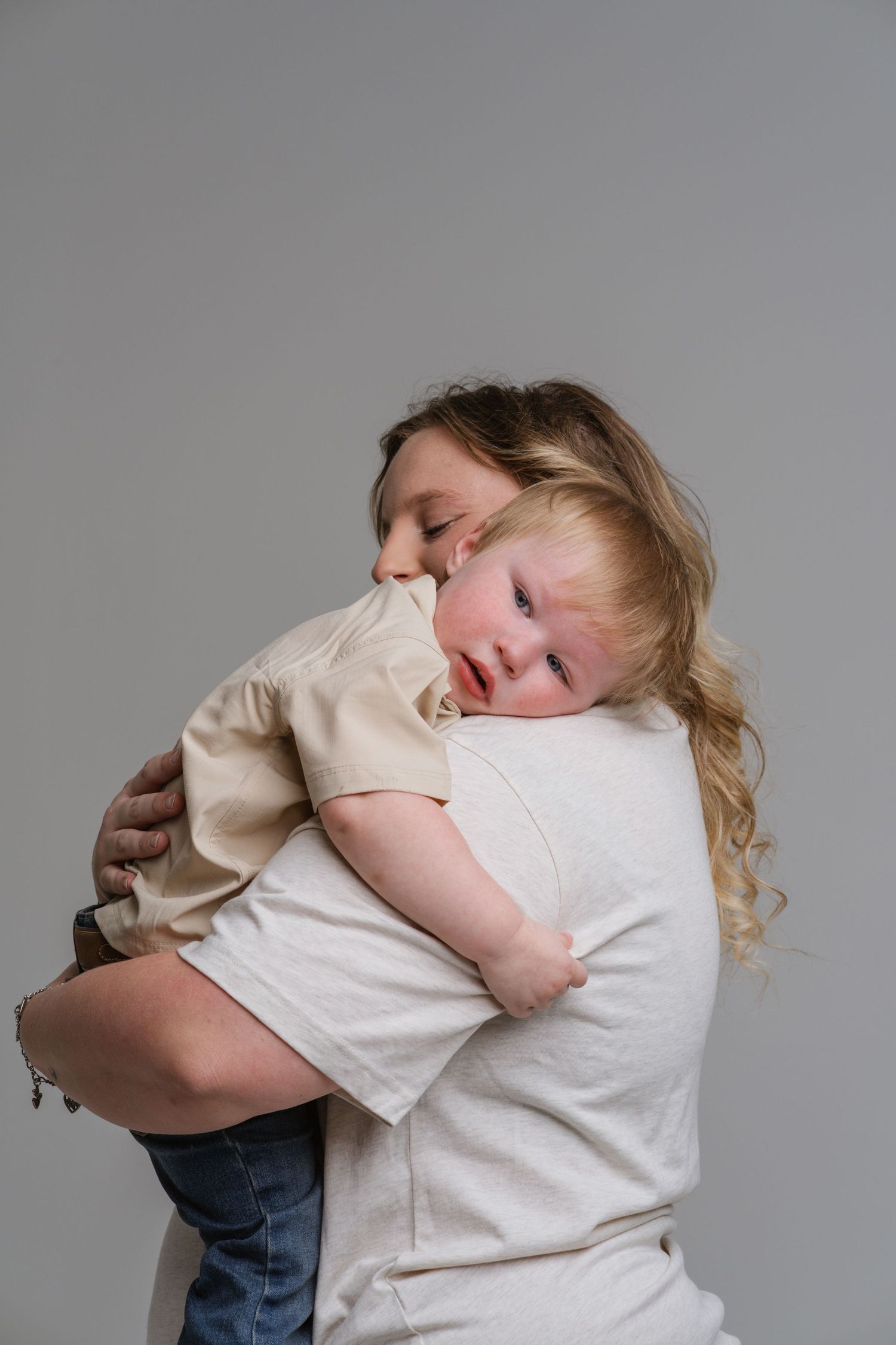 A mother with curly blonde hair holds her young toddler close in a warm embrace against a gray background.