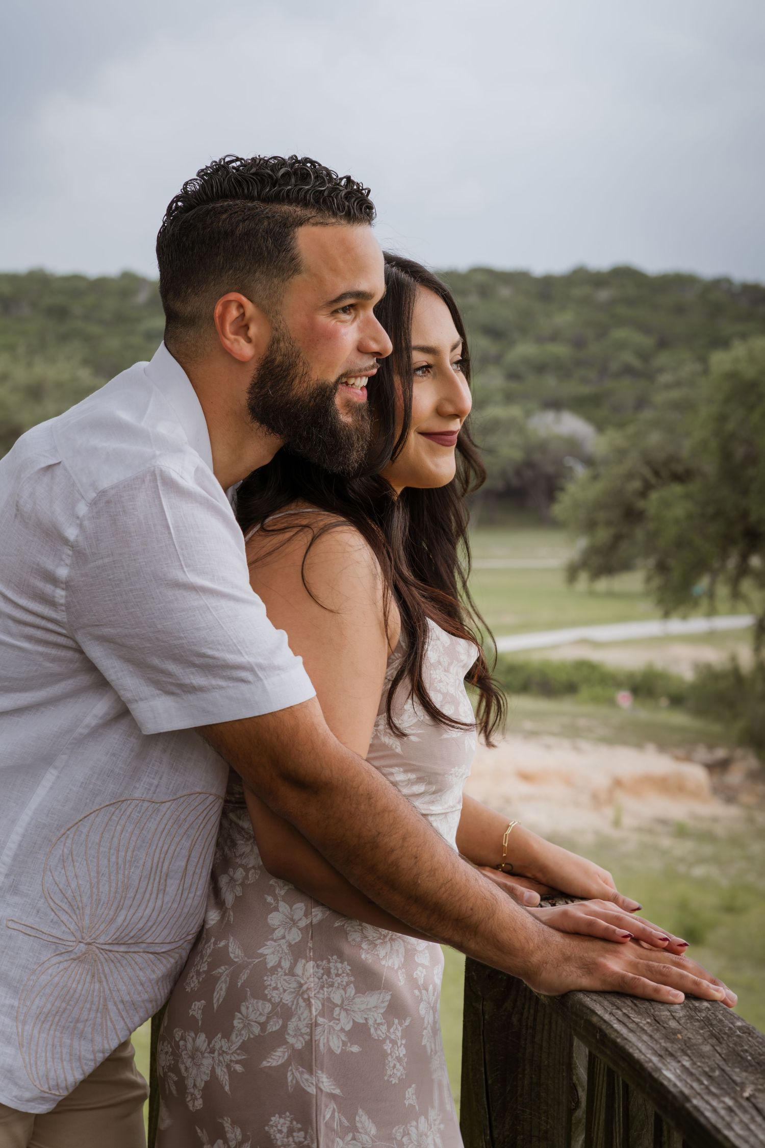 A couple leans on a wooden railing, smiling as they gaze at a lush green landscape on a cloudy day.