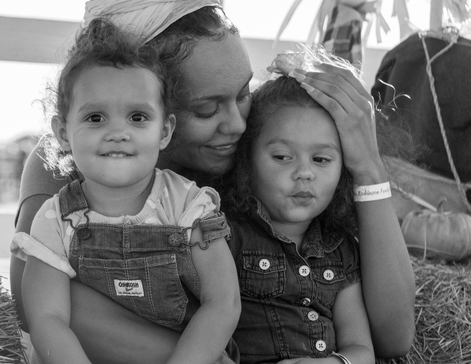 Candid family portrait of a mother and two young children sitting together outdoors at a pumpkin patch.