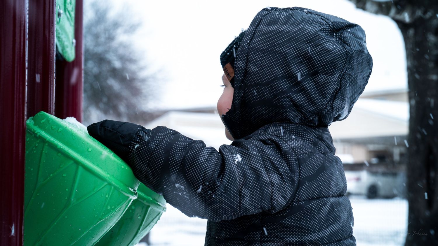 Child in a winter coat placing an item into a green bin outdoors during a snowy day.