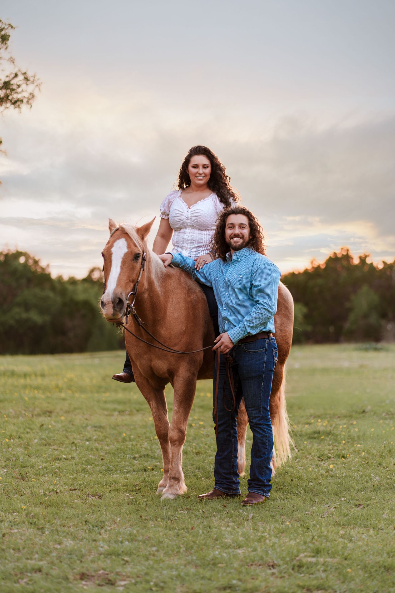 Couple posing with a chestnut horse in a green field at sunset, woman sitting on horseback, man standing beside.