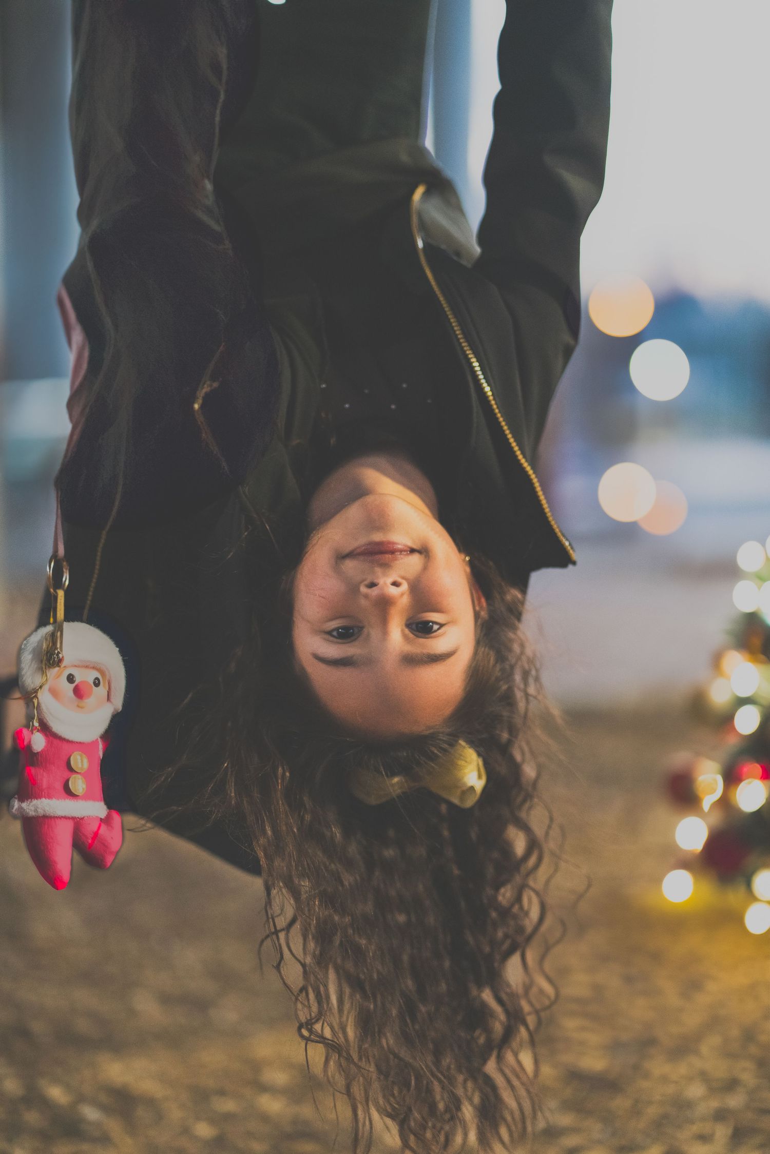 A person hanging upside down with long curly hair and a Santa keychain dangling against a blurred bokeh background.
