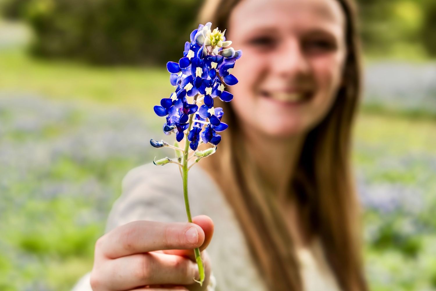 Close-up of a hand holding a bluebonnet flower with a smiling person softly blurred in the background.