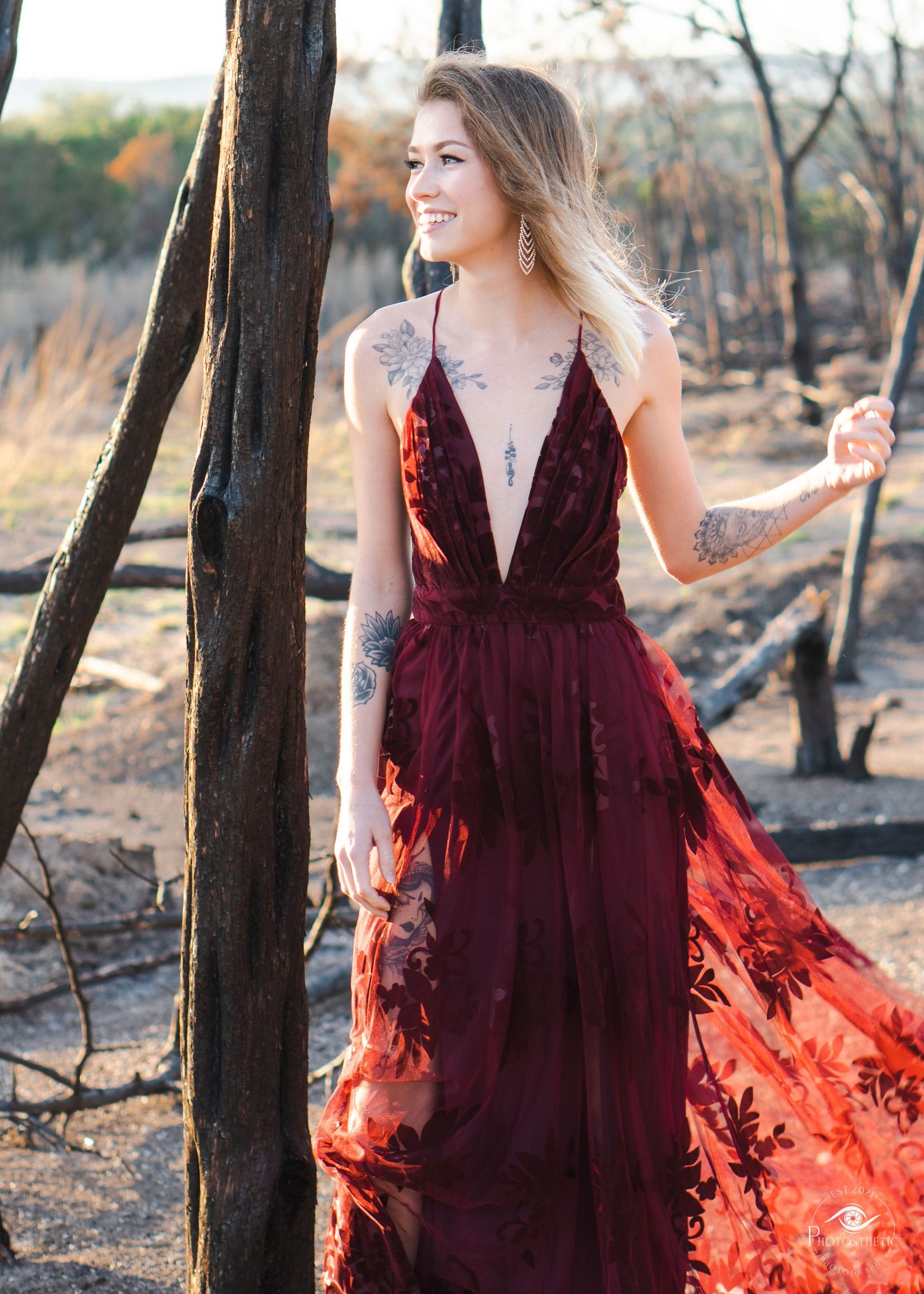Artistic outdoor portrait of a tattooed woman in a flowing red dress, captured in soft natural light.